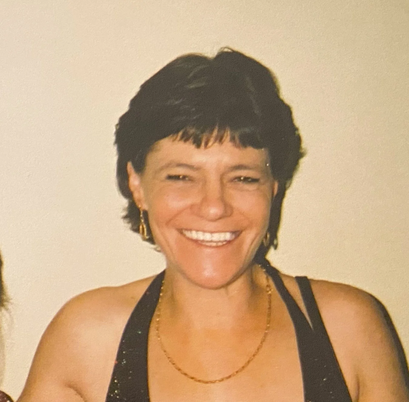 Photo of a woman with short dark hair, smiling, wearing earrings, a gold necklace, and a black top with thin straps, against a plain light background.