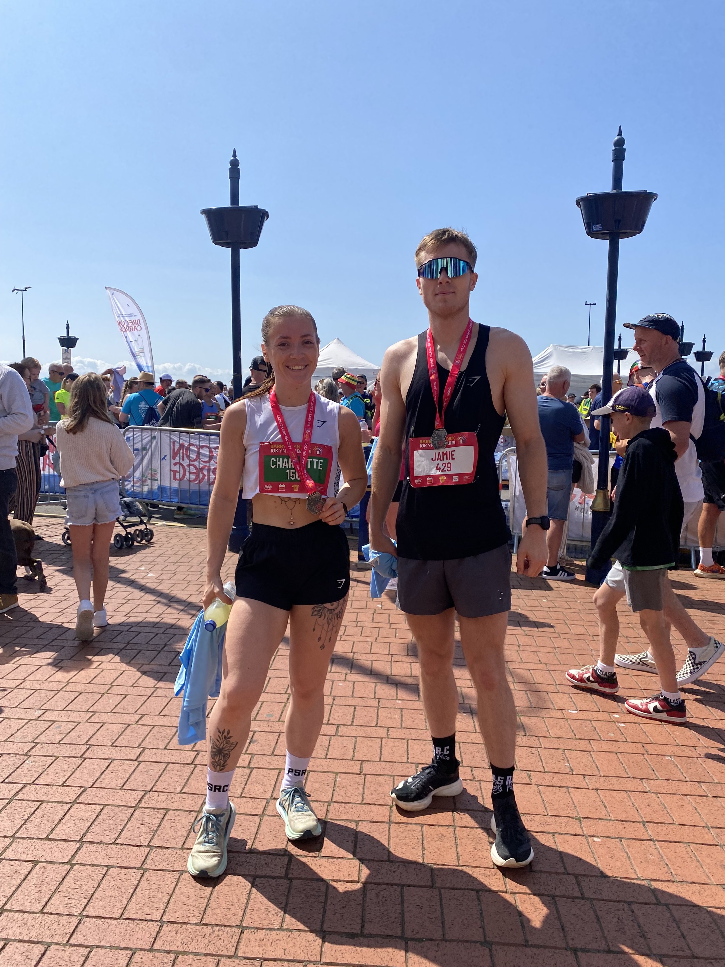 Two marathon runners, a woman and a man, standing together after finishing a race, wearing medals and race bibs, with a crowd of people and event tents in the background.