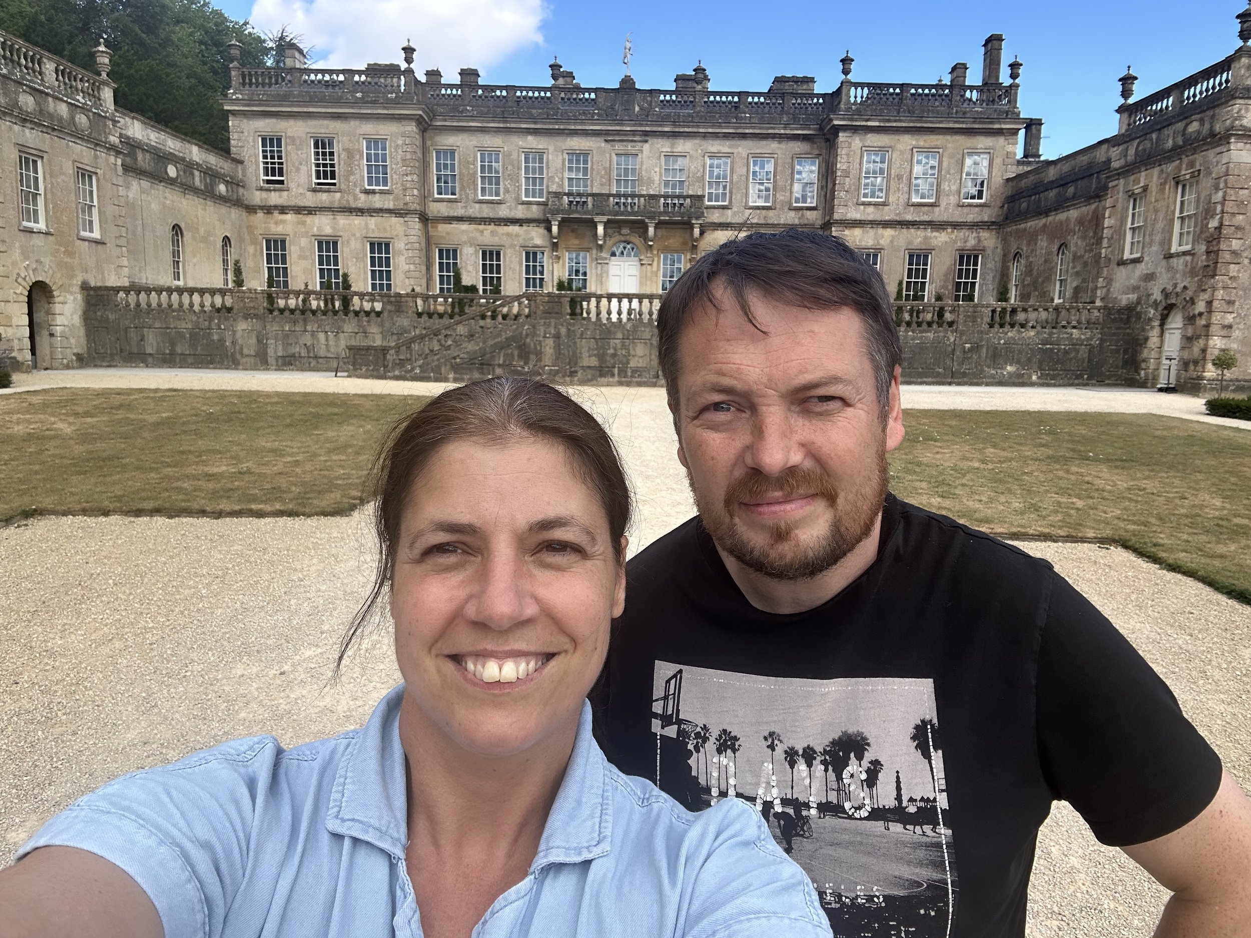 A selfie of a woman and a man in front of a historic mansion with multiple windows, stone walls, and decorative stoneposts along the roofline.