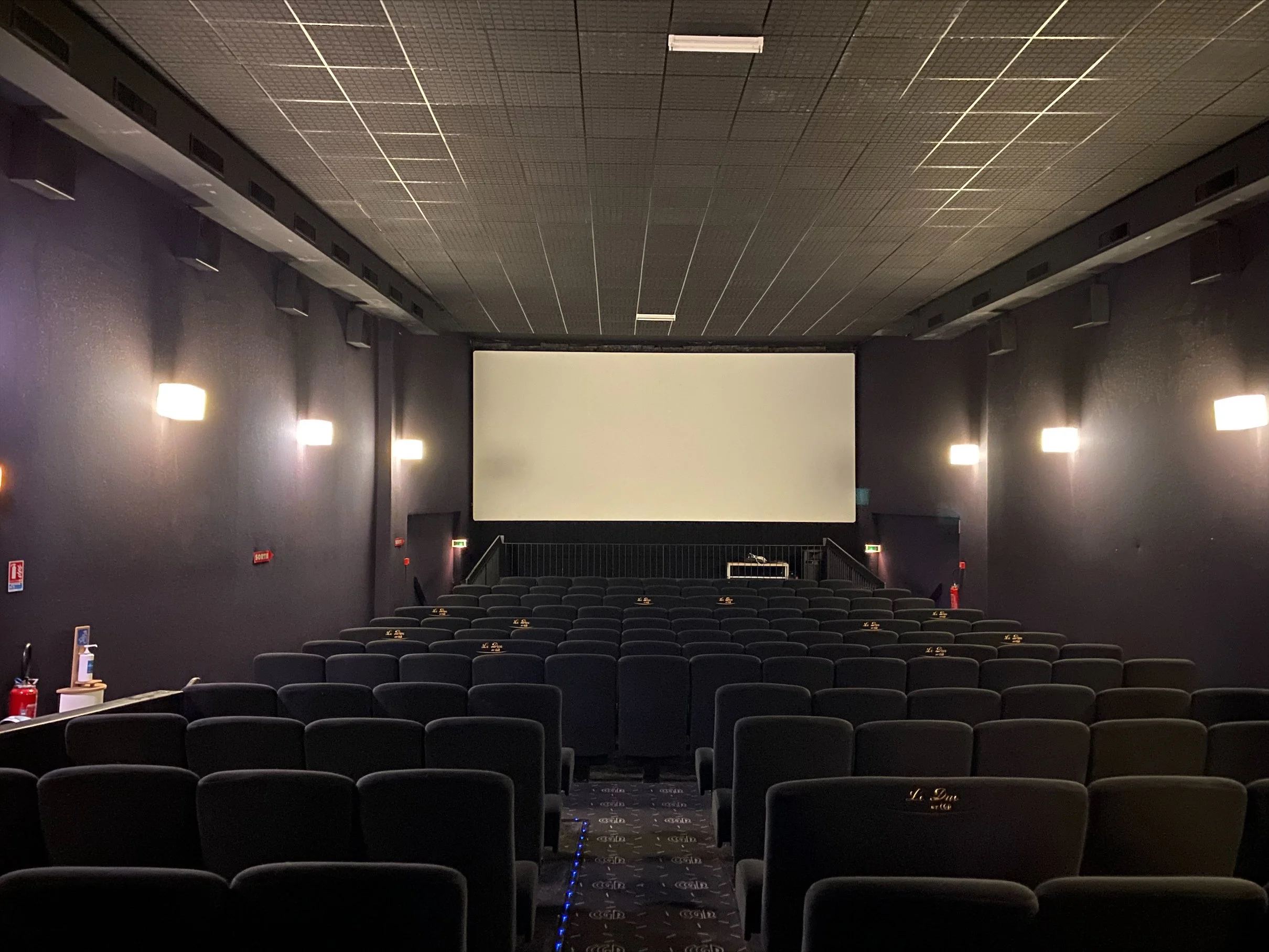 Image d'une salle de cinéma du CGR avec des rangées de fauteuils noirs, un grand écran blanc à l'avant, des murs violets et des lumières murales brillantes.