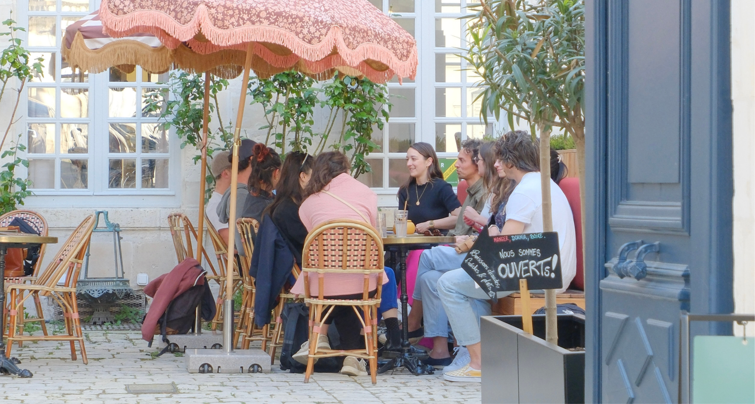 Groupe de personnes assises à une table extérieure dans un café, discutant et souriant sous un parasol rose, avec des plantes vertes autour et un panneau indiquant que le lieu est ouvert.