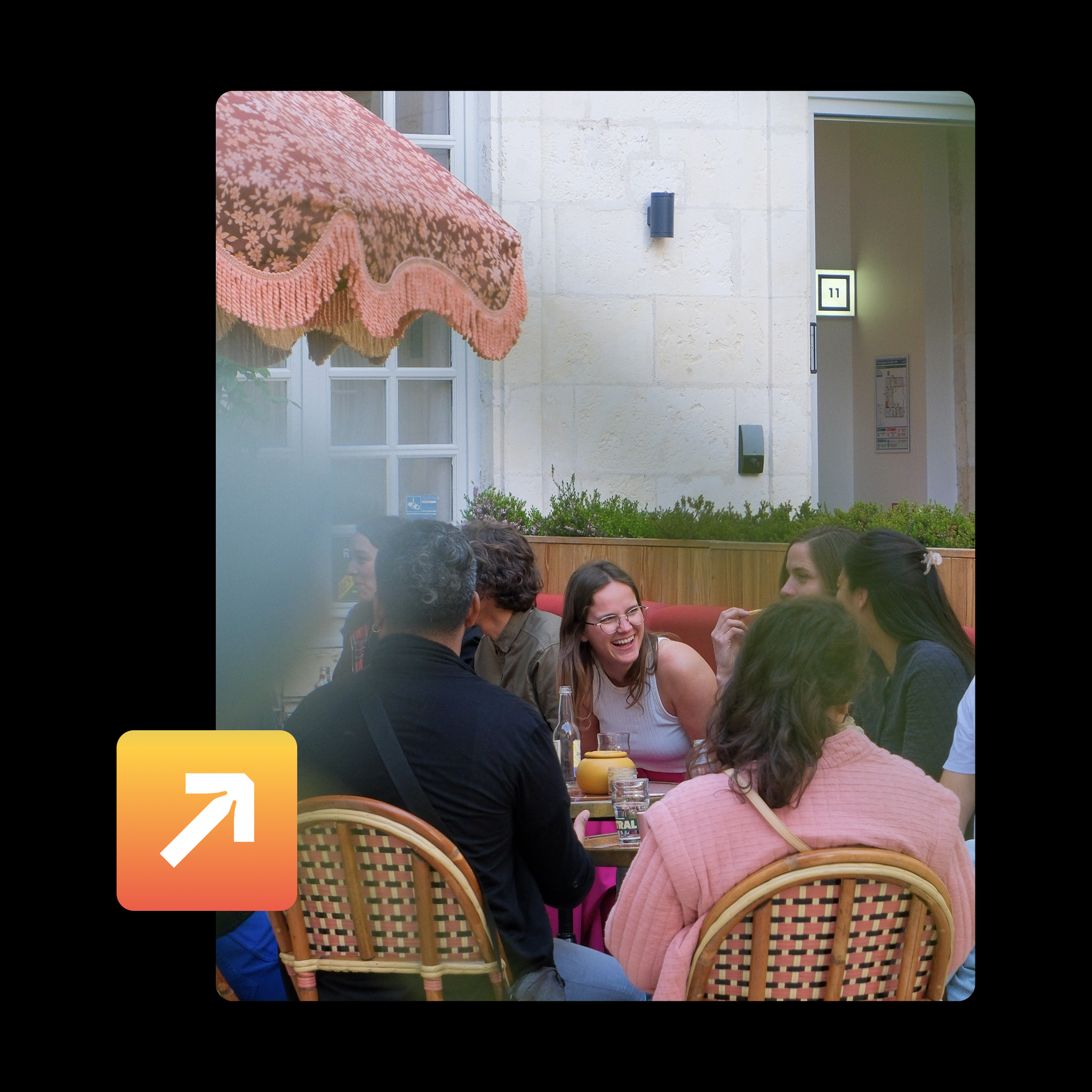 Groupe de personnes assises autour d'une table, riant et discutant dans un restaurant ou un café, avec des chaises en rotin et des décorations en arrière-plan.