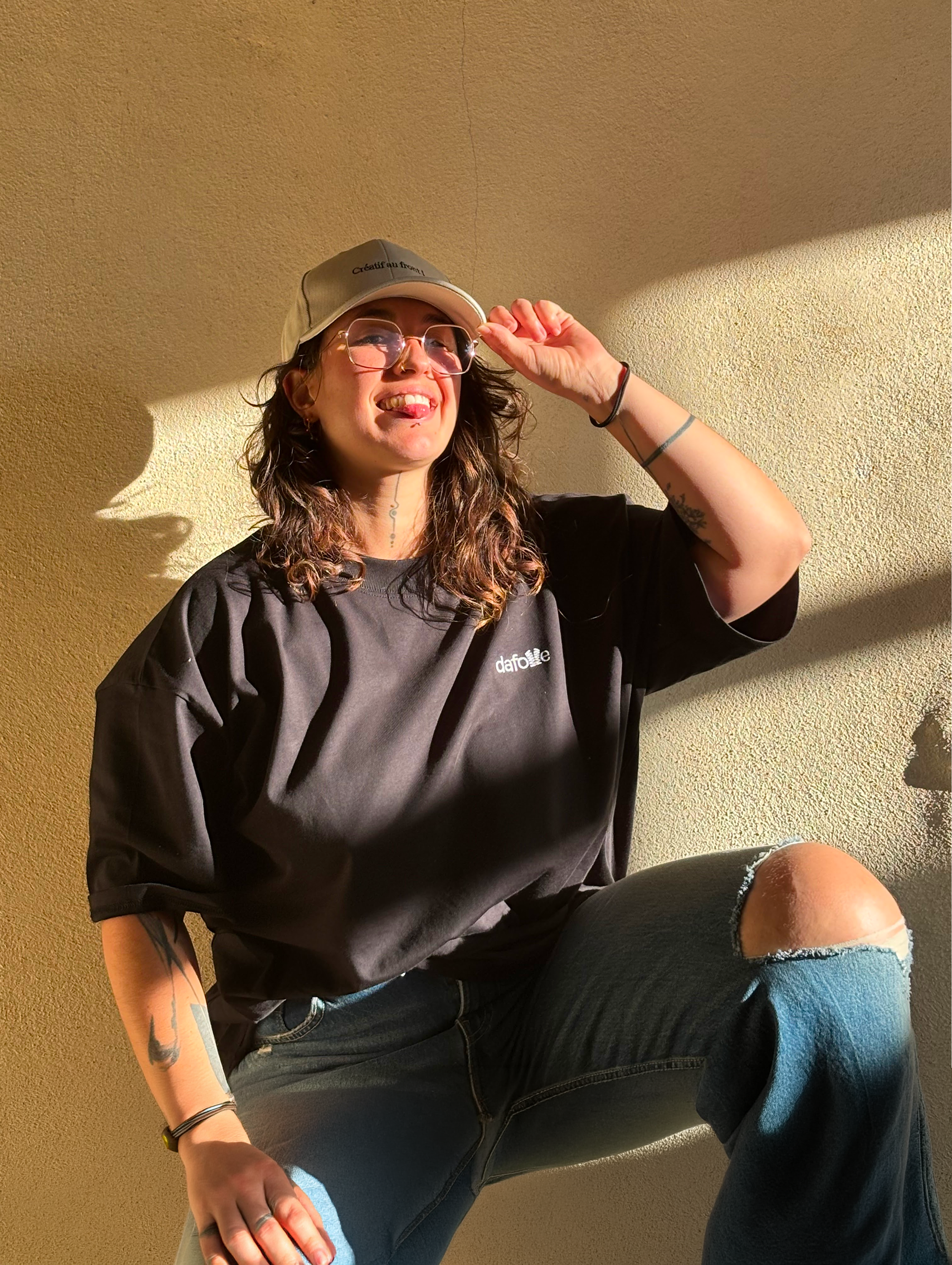 Lili Schmitt, Secrétaire générale de Keyframers Collective. Un tatouage au bras et des bracelets, assise sur le sol, souriante avec la langue sortie, portant un sweat noir avec le logo dafolle et un jean déchiré, dans une pièce avec un mur beige.