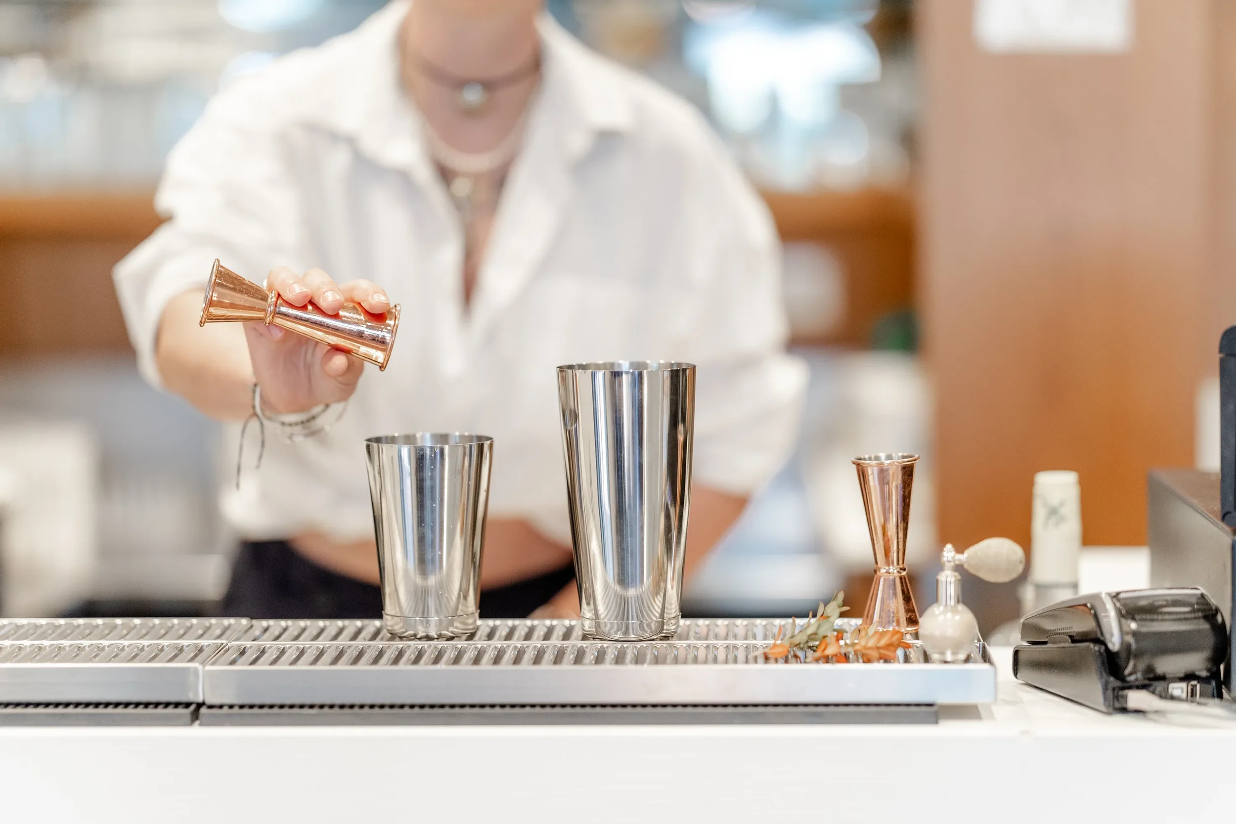 Une personne verse une boisson dans un verre en métal dans un bar ou un restaurant, avec plusieurs autres verres et ustensiles sur le comptoir.