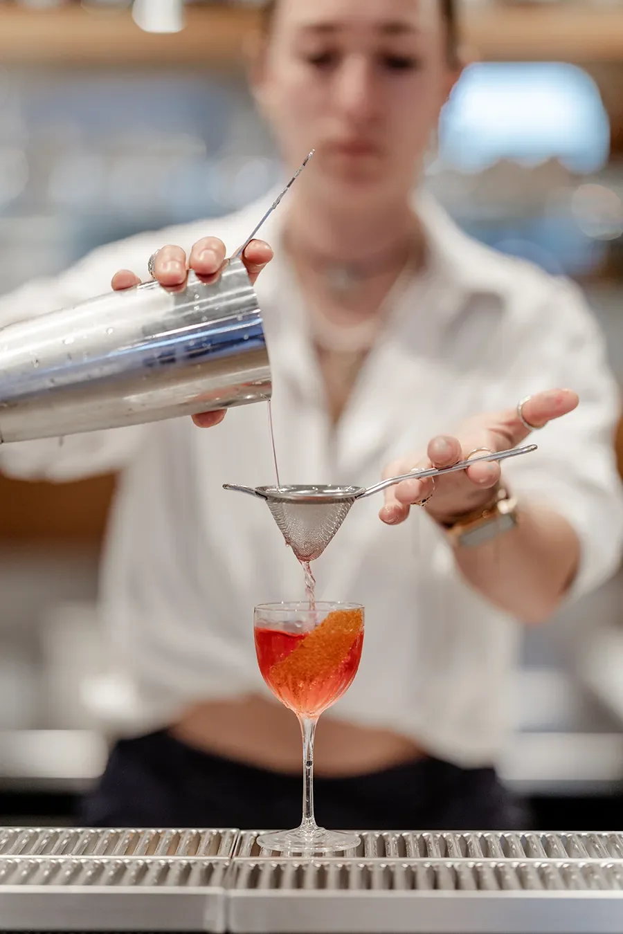 Un chef ou barman filtre un cocktail rouge dans un verre à vin à l'aide d'une passoire fine.