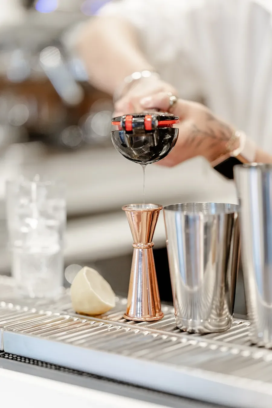 Un barman verse du liquide noir dans une tasse à cocktail en utilisant un jigger en cuivre, avec des outils de bar en métal à proximité