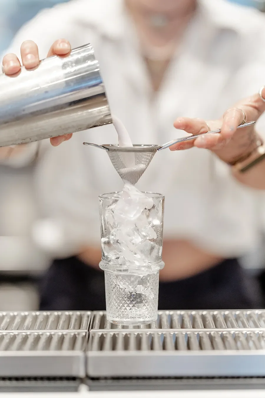Une personne verse un liquide blanc à travers un tamis dans un verre rempli de glaçons.