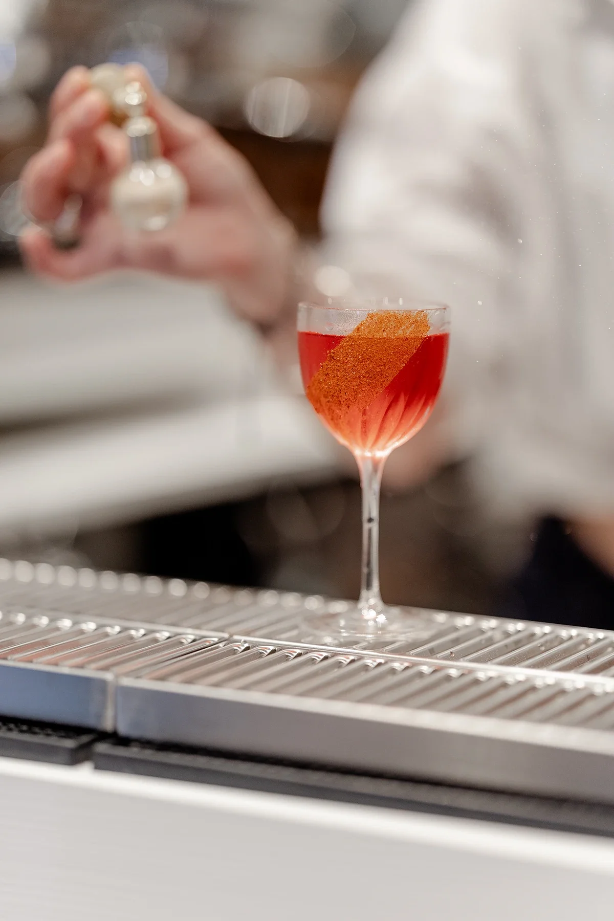 Un verre à cocktail avec une boisson rouge, posé sur un bar, avec une personne en arrière-plan utilisant un spray à vaporiser.