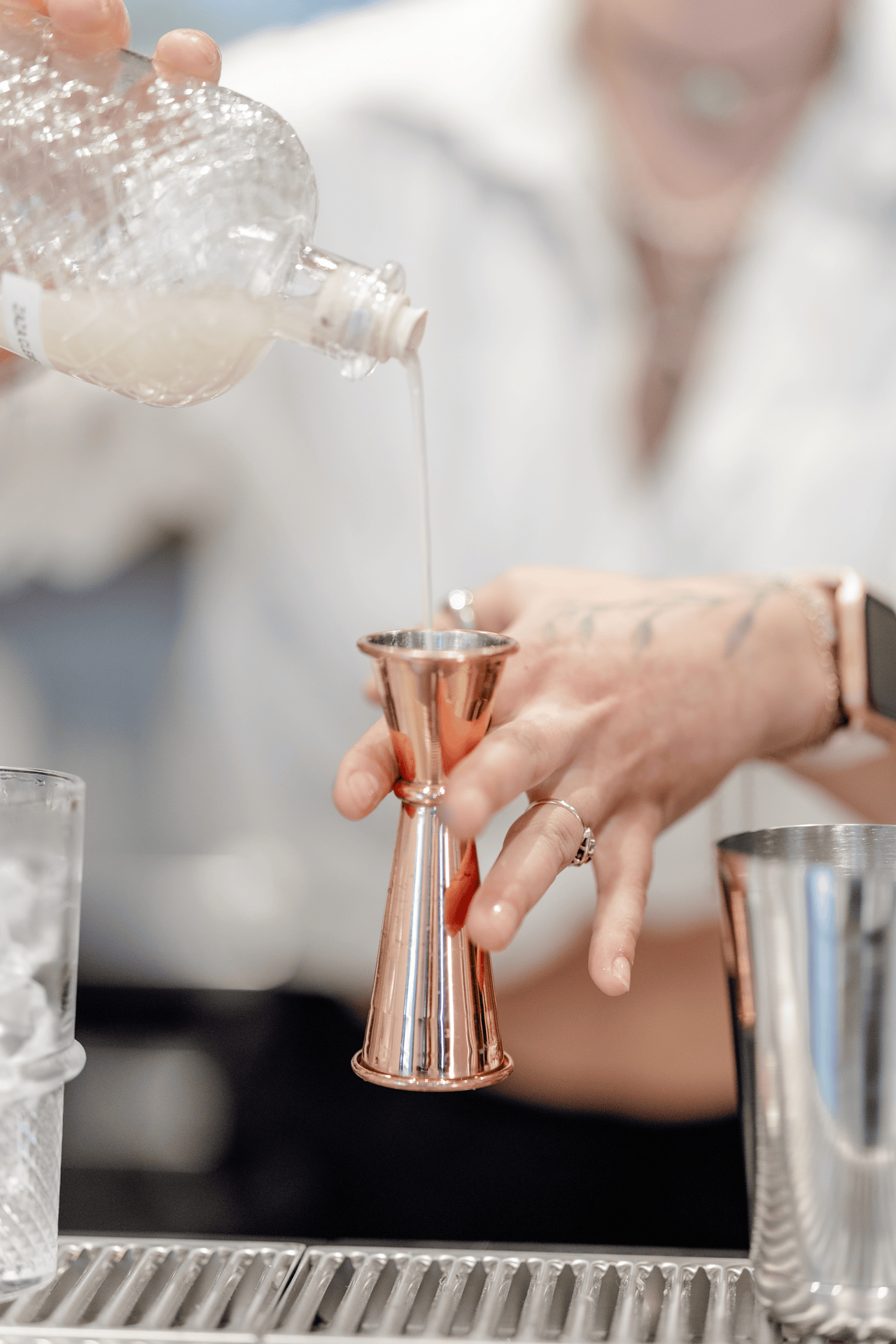 Personne préparant un cocktail dans un bar avec une bouteille de liquide transparent, un jigger en cuivre, et divers ustensiles de bar sur le comptoir.