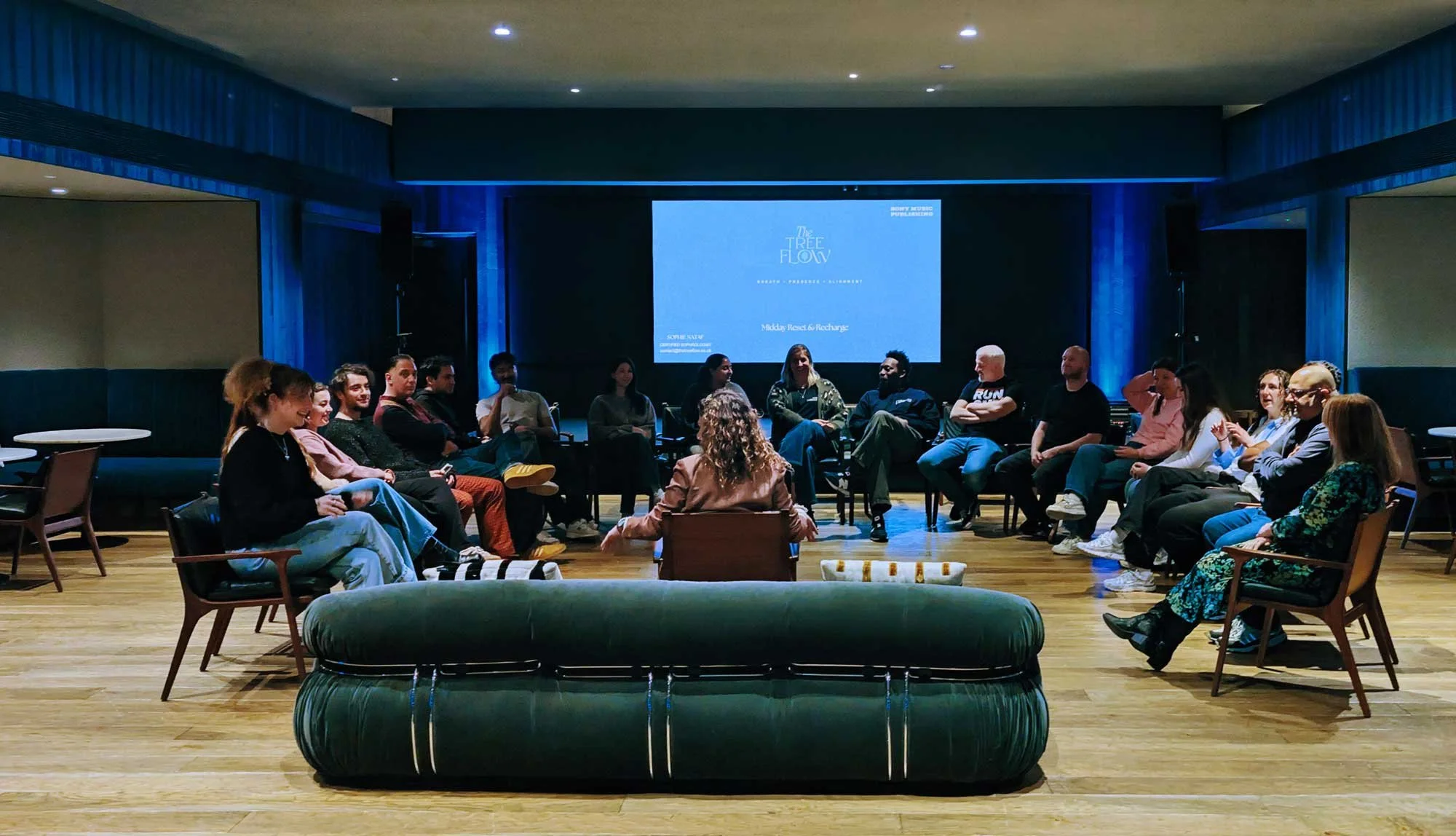A group of people seated in a circle in a dimly lit room, participating in a discussion or workshop, with a large screen displaying a presentation titled 'The True Flow' in the background.