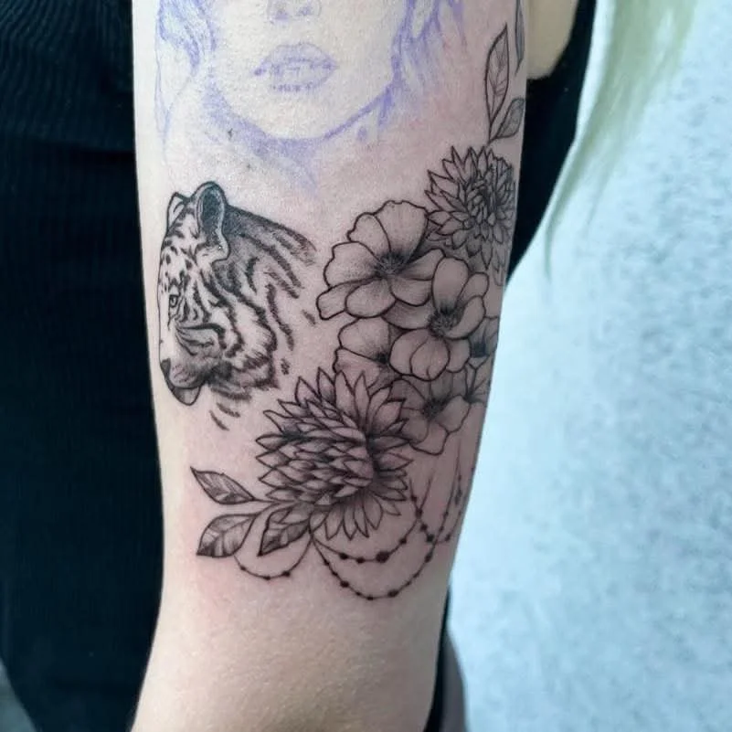 Tattoo auf dem Arm mit einem Tigerkopf, Blumen und Blättern.