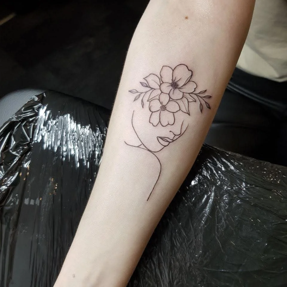 Tattoo eines minimalistischen Gesichts mit Blumen und Blättern auf der Stirn.