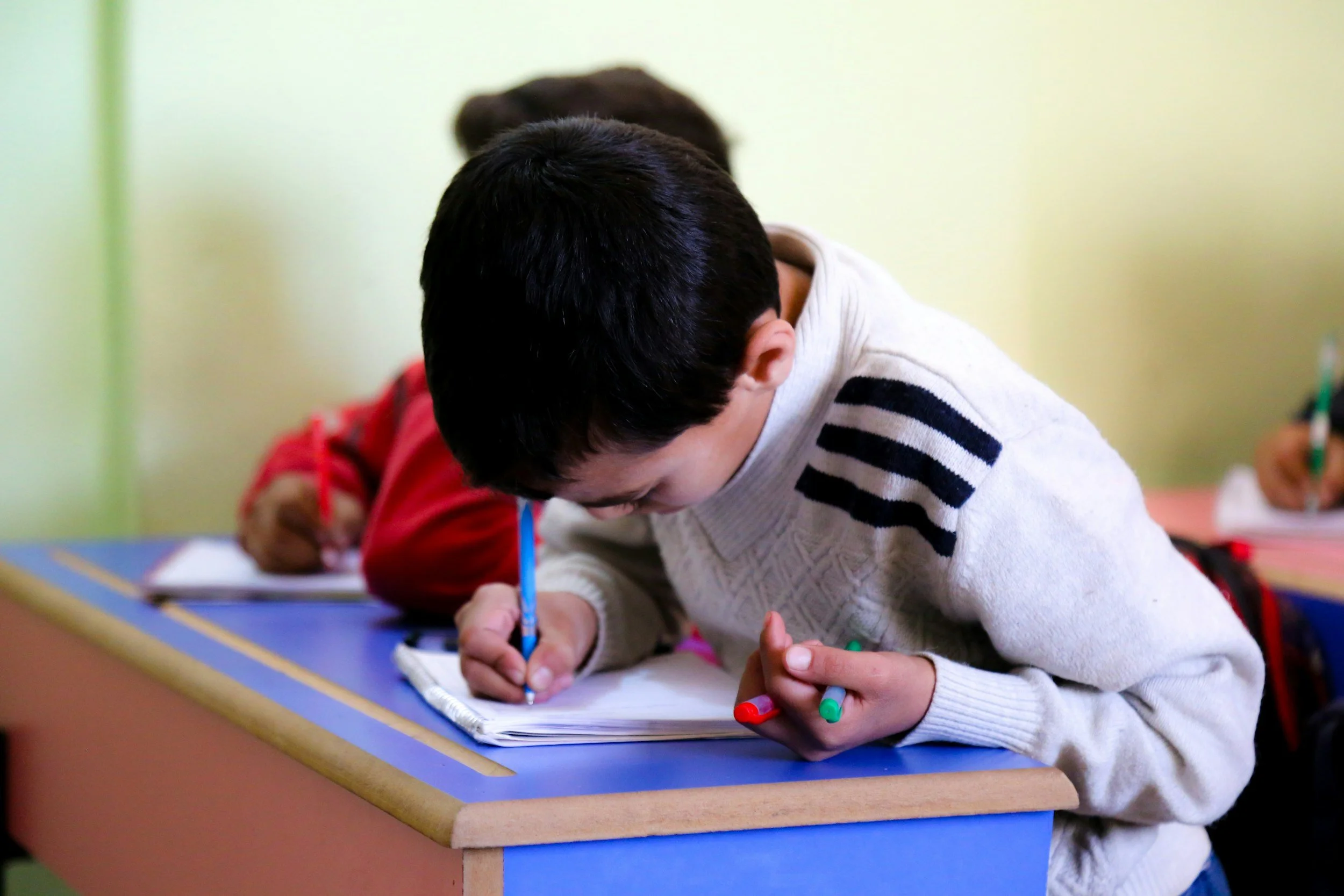 Children sitting at a table, writing or drawing on paper with colored markers in a classroom setting.