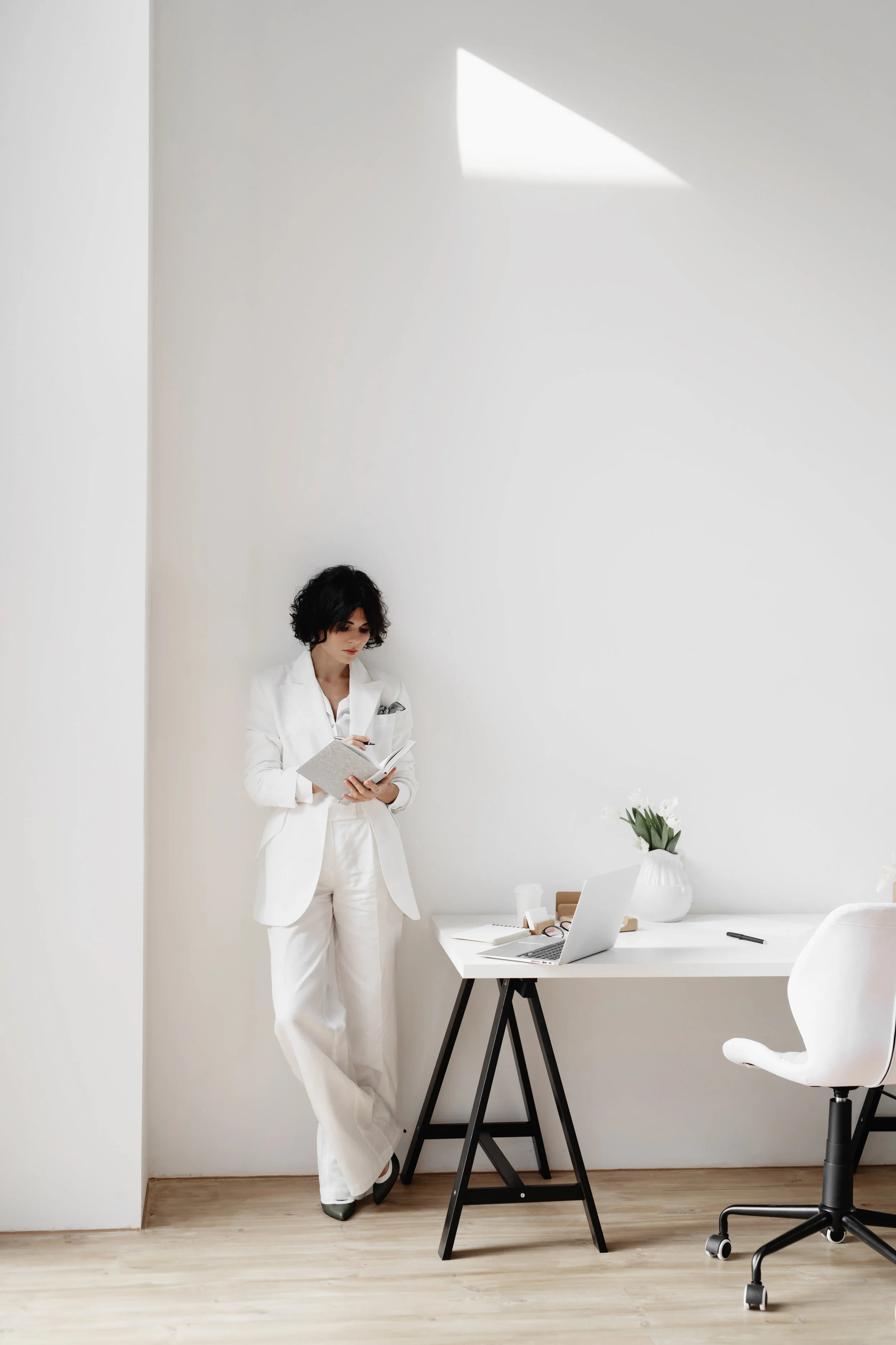 A woman dressed in a white suit, standing by a white wall, reading a notebook in a minimalistic office with a white desk, a silver laptop, a white vase with flowers, and an office chair.