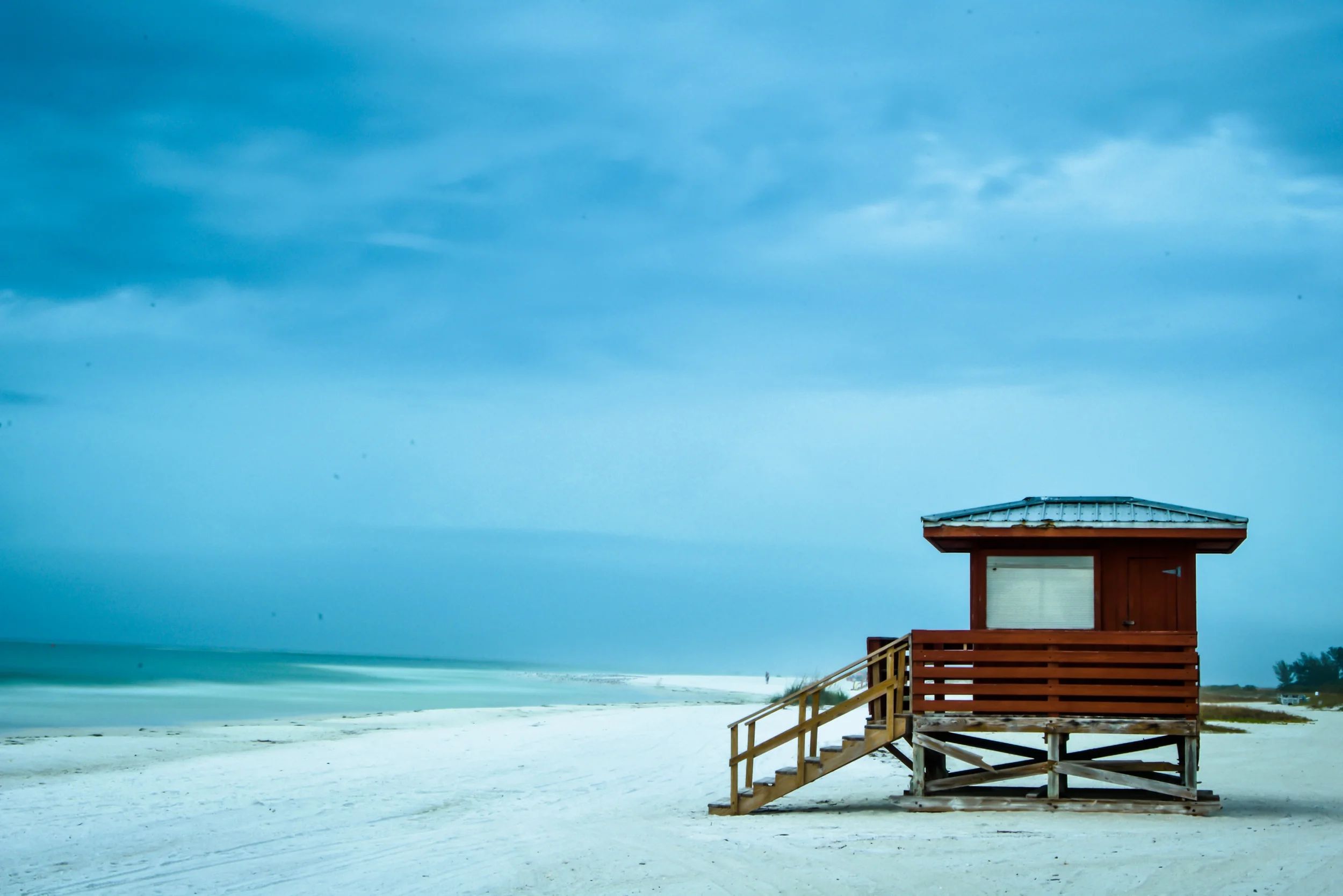 Sarasota Motor Club - Premium Car Service Sarasota - A wooden beach lifeguard tower on a sandy shore, with an overcast sky and the ocean in the background.