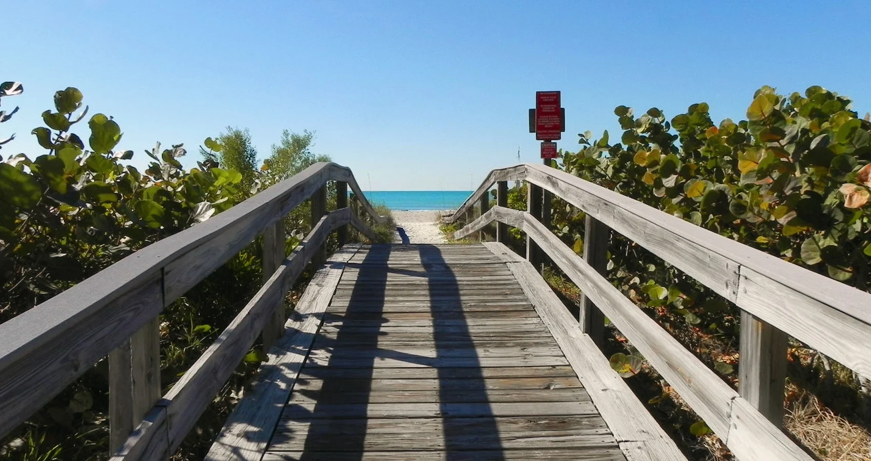 Sarasota Motor Club - Exotic Car Consignment Sarasota - Wooden boardwalk leading to a beach with clear blue sky and ocean in the background, surrounded by green shrubbery and with red signs at the end.