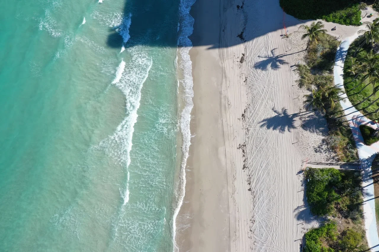 Sarasota Motor Club - Exotic Car Consignment Sarasota - Aerial view of a tropical beach with turquoise water, white sand, palm trees, and shadows cast by the trees.