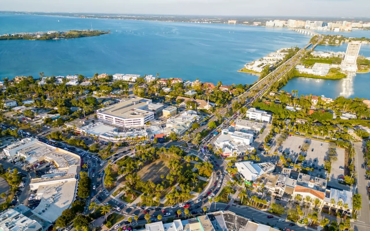 Sarasota Motor Club - Sarasota Luxury Car Rental - Aerial view of a coastal city with water, a bridge, buildings, roads, and green trees.