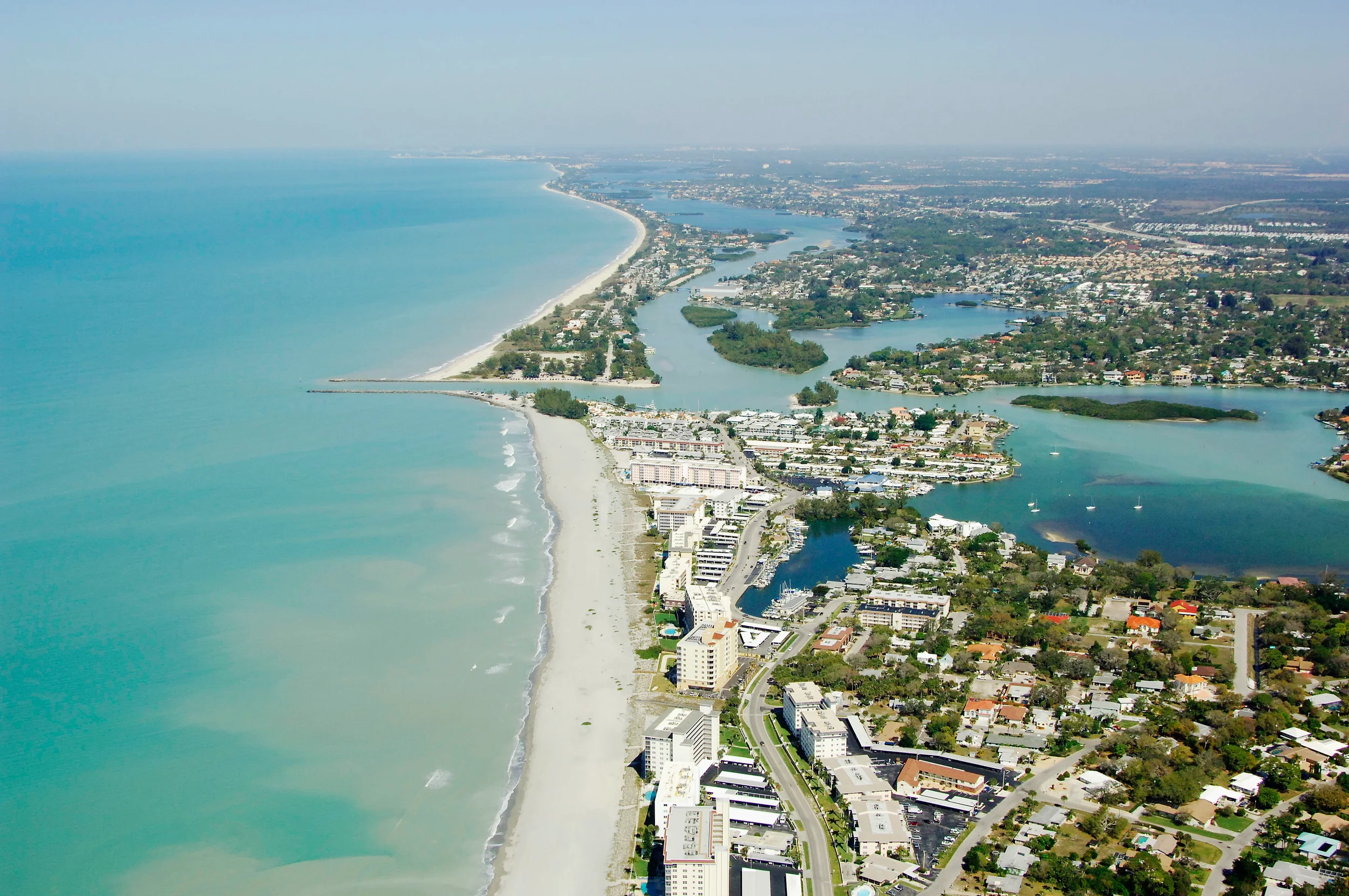 Sarasota Motor Club - Exotic Car Consignment Sarasota - Aerial view of a coastal city with a long sandy beach, ocean, and inland waterways surrounded by residential and commercial buildings.