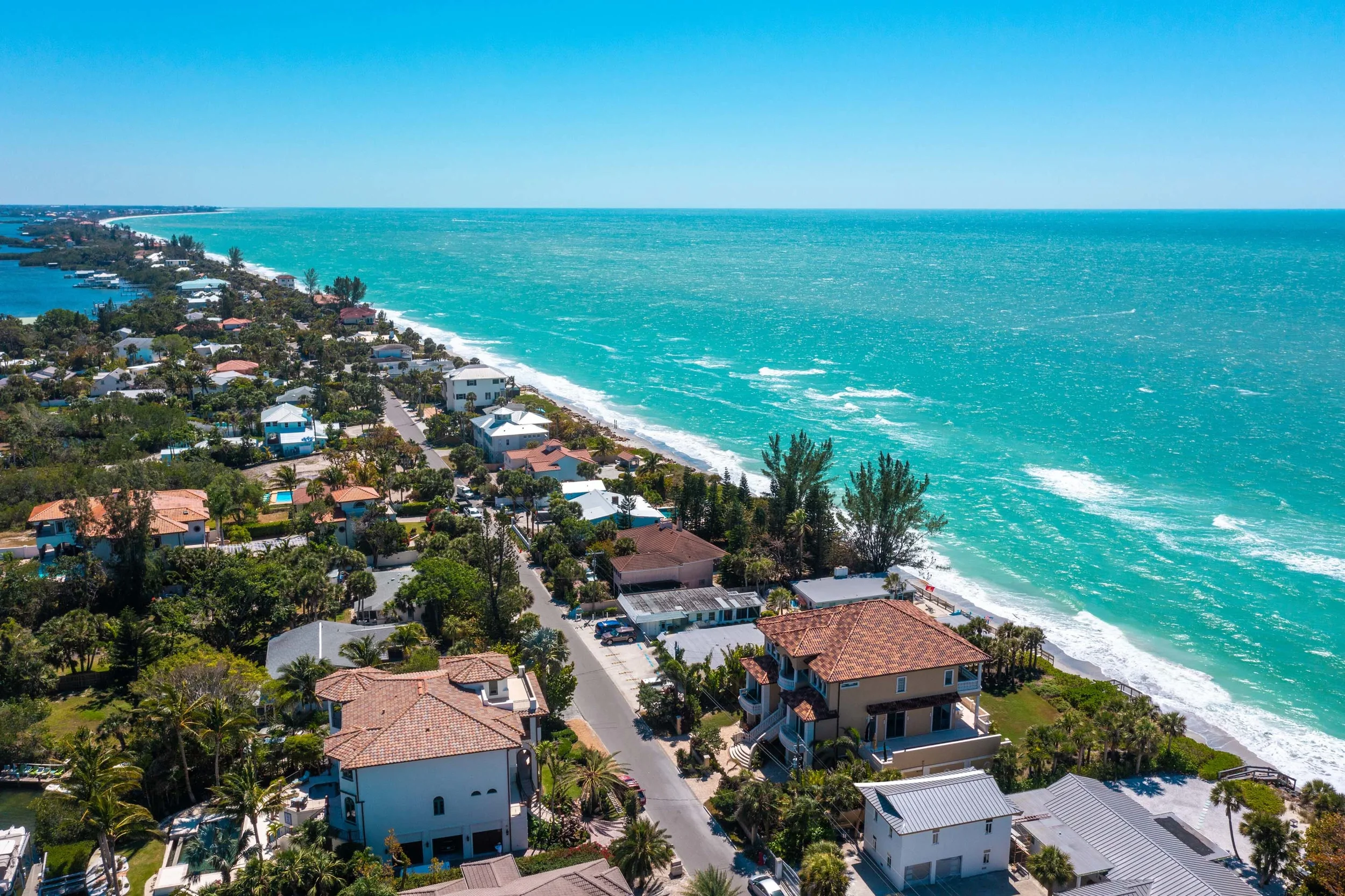 Sarasota Motor Club - Exotic Car Consignment Sarasota - Aerial view of a coastal neighborhood with houses, palm trees, and a beach along the turquoise ocean under a clear blue sky.