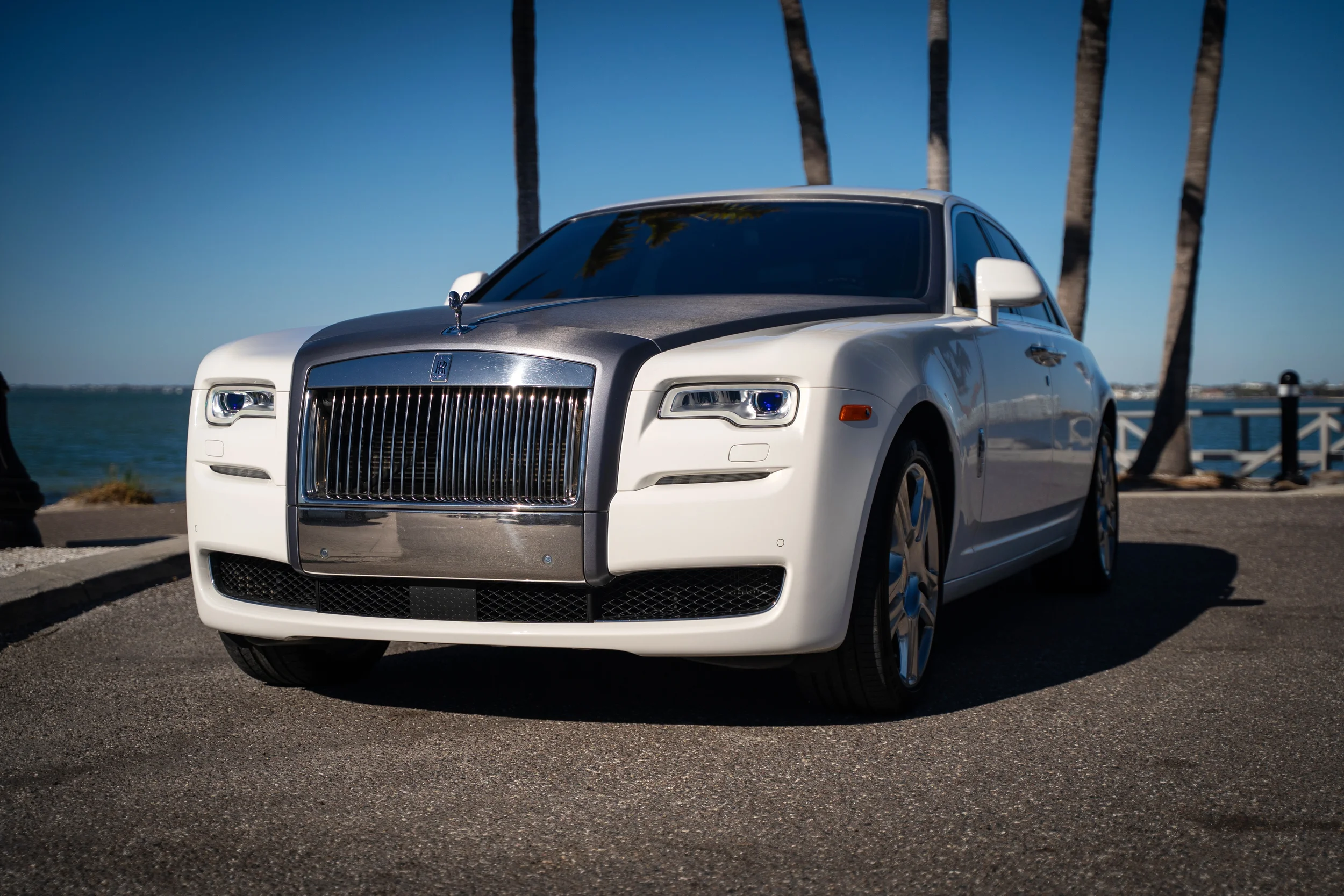 A white luxury Rolls-Royce car parked near a waterfront with trees and a clear blue sky in the background.