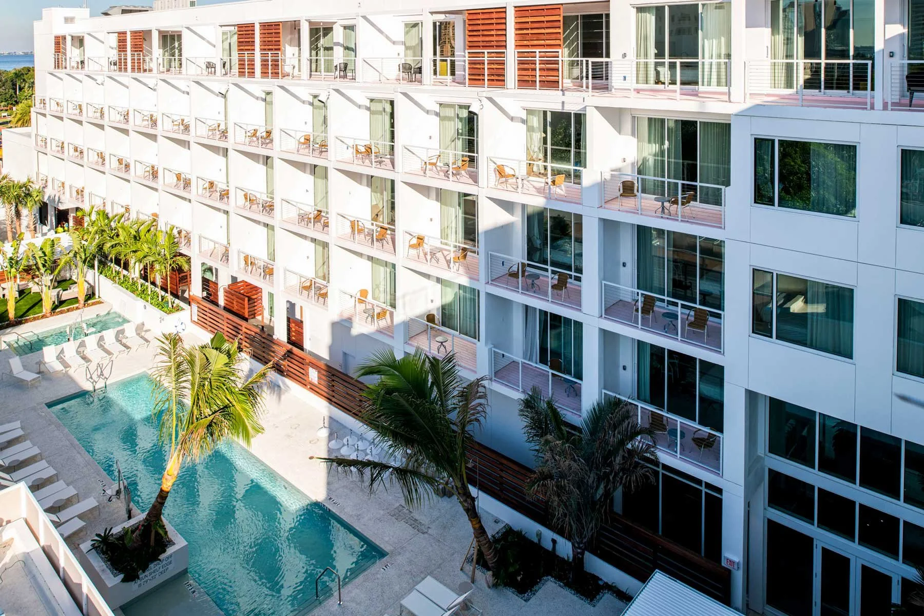 View of a modern hotel with multiple balconies, a swimming pool, and palm trees in a tropical or coastal setting.