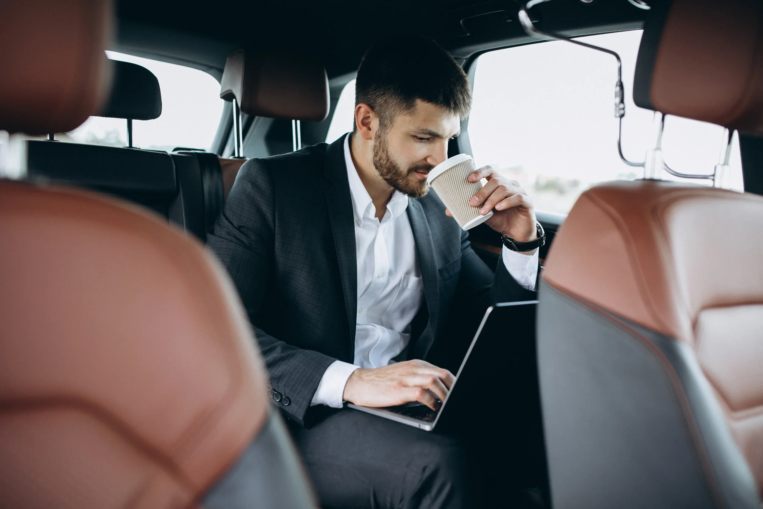 Sarasota Motor Club - Exotic Car Rental Sarasota - Man in business suit sitting in back of car using a laptop, holding a coffee cup, with brown leather seats.