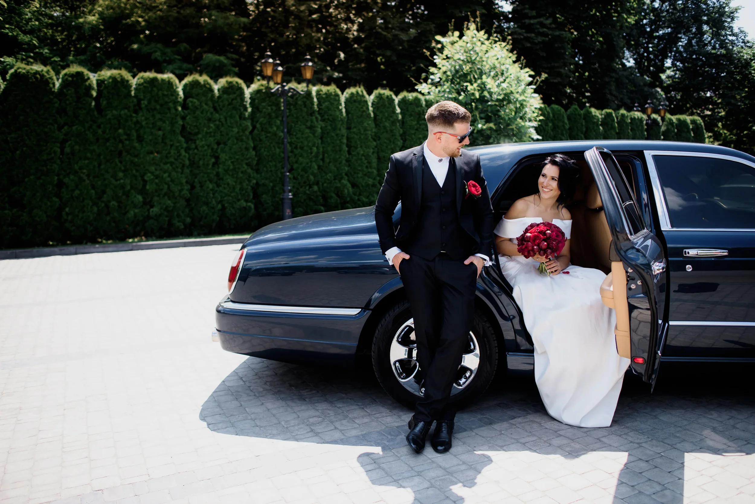 Sarasota Motor Club - Exotic Car Rental Sarasota - A newlywed couple with the groom in a black suit and the bride in a white wedding dress, posing near a black luxury car outdoors with green trees and bushes in the background on a sunny day.