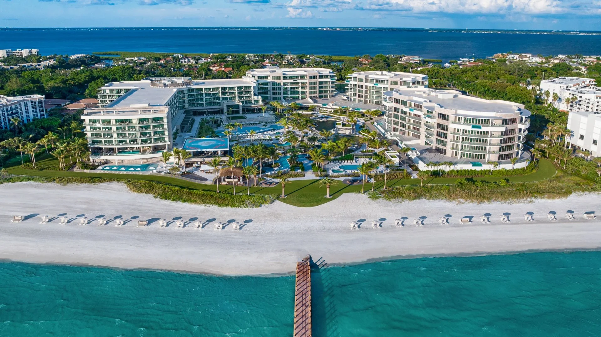 Sarasota Motor Club - Premium Car Service Sarasota - Aerial view of a beachfront resort with multiple modern white buildings, swimming pools, palm trees, and a sandy beach facing the ocean.