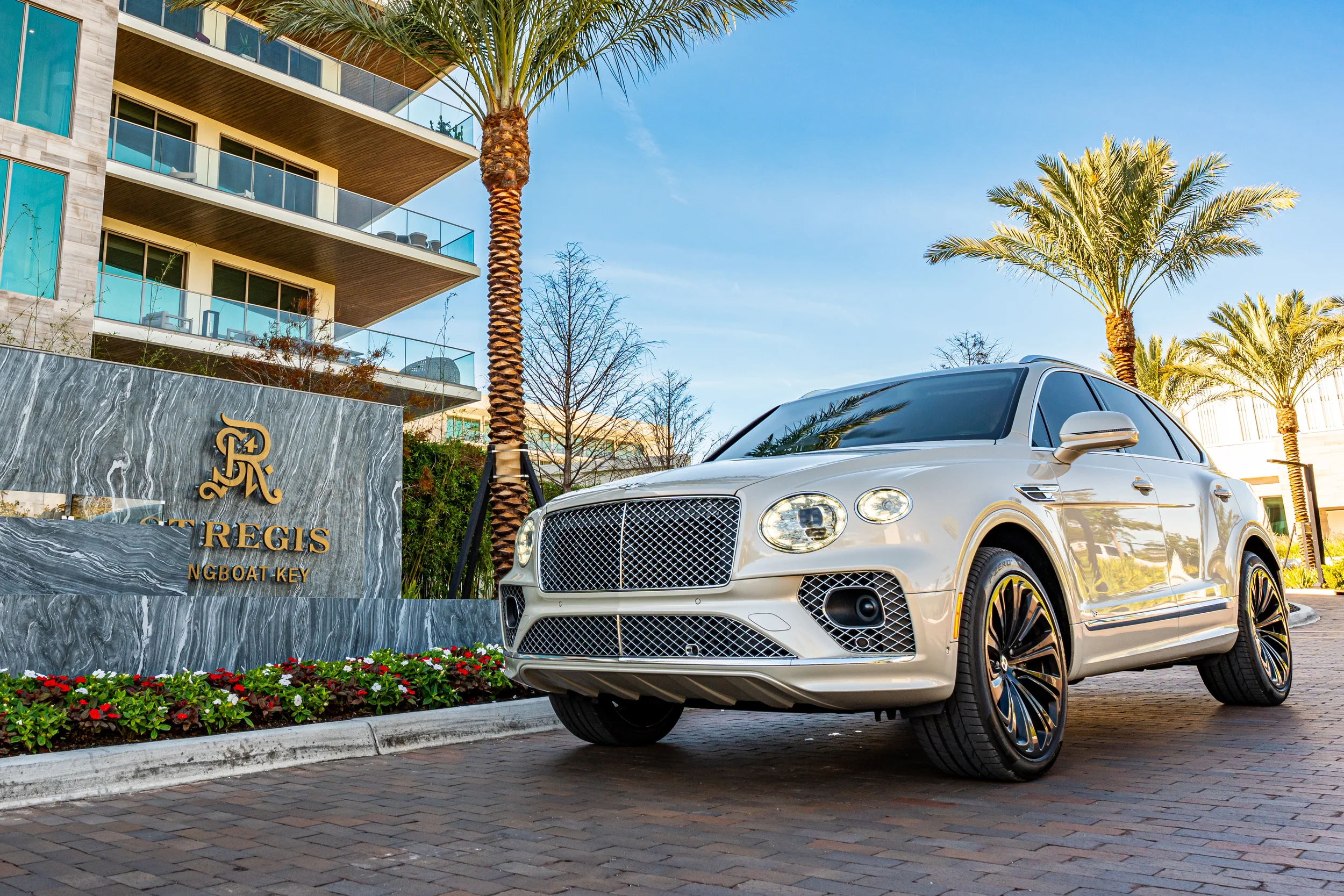 Sarasota Motor Club - St. Regis luxury car rental - A white luxury Bentley Bentayga  parked in front of a modern apartment building with palm trees and a stone sign that reads 'The Residences at Regale Ngboat Key'.