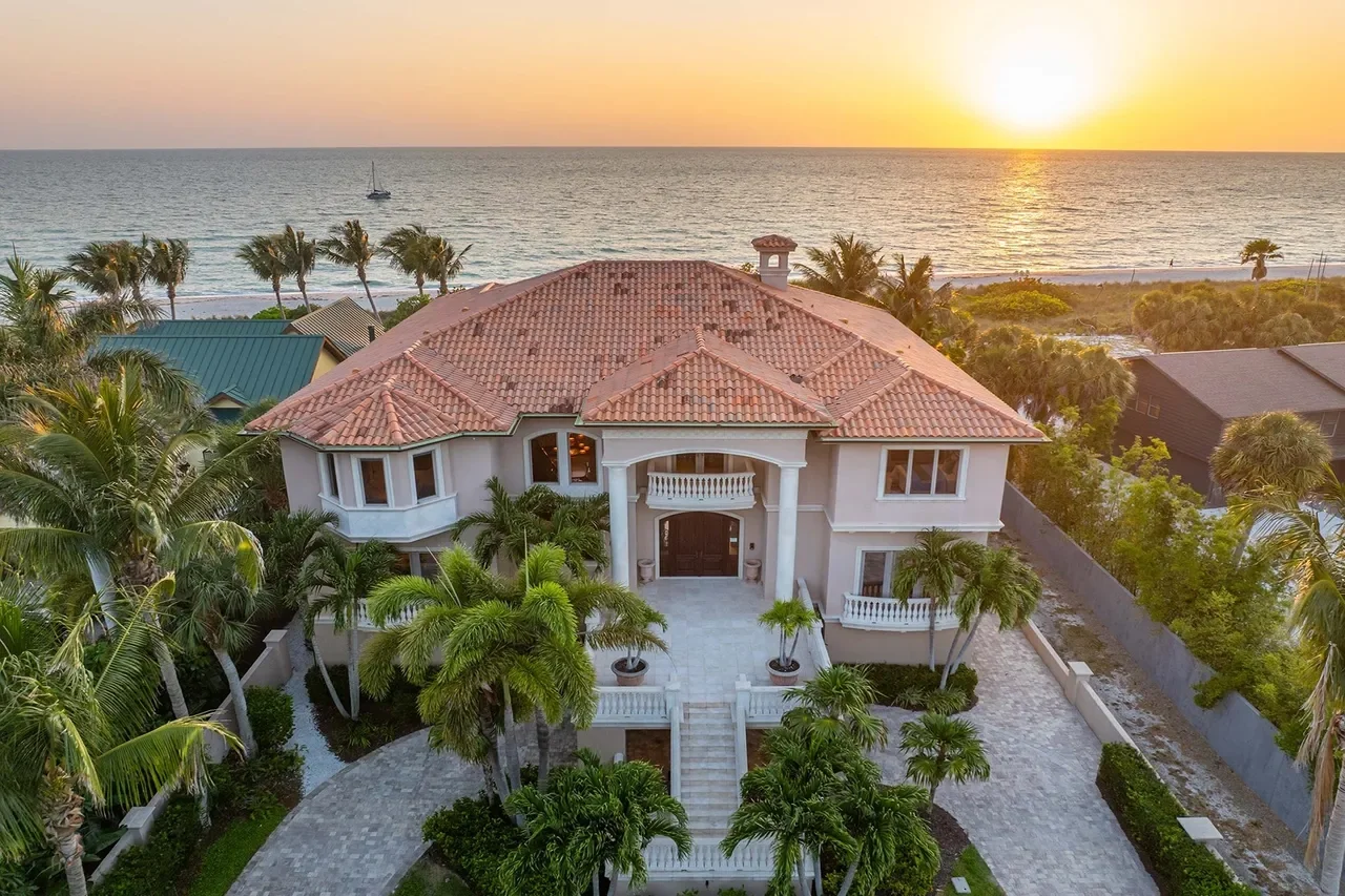 Sarasota Motor Club - Exotic Car Consignment Sarasota - A large beachfront house with a red tile roof, surrounded by palm trees, overlooking the ocean at sunset.