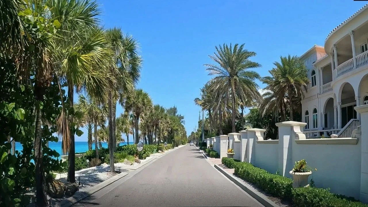 Sarasota Motor Club - Exotic Car Consignment Sarasota - A scenic street lined with palm trees on one side and white buildings with arches on the other, with a view of the ocean in the background under a clear blue sky.
