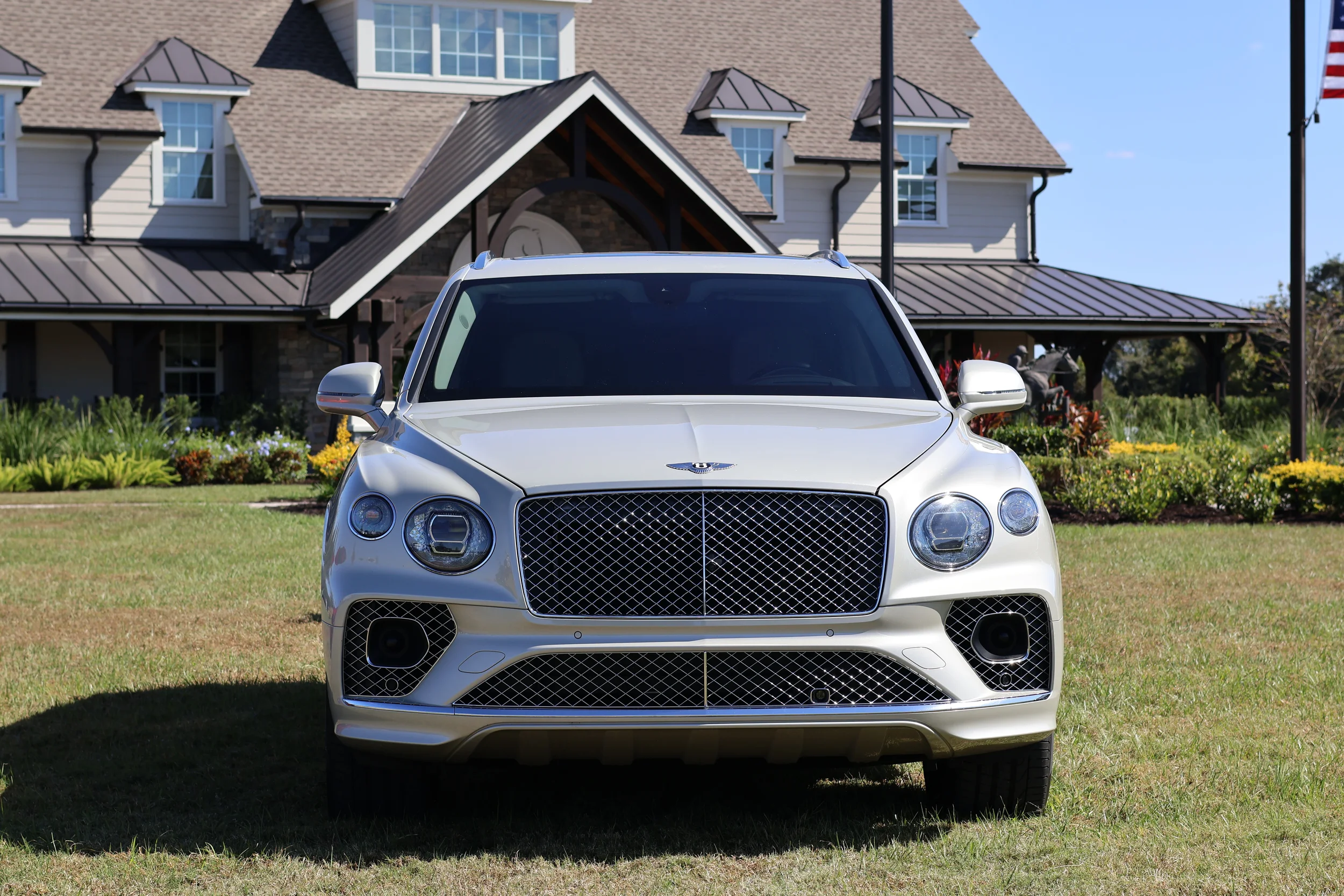 Sarasota Motor Club - Exotic Car Consignment Sarasota - Front view of a white Bentley parked on grass in front of a large house with a manicured lawn, flower beds, and American flags.