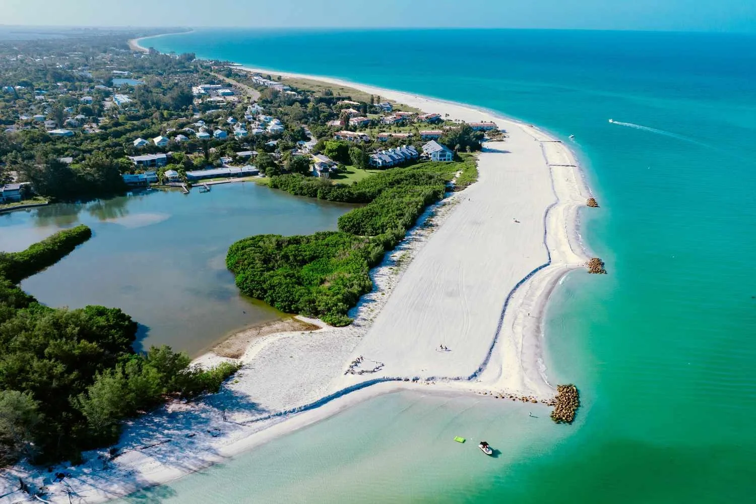 Sarasota Motor Club - Premium Car Service Sarasota - Aerial view of a coastal area with a sandy beach extending into the ocean, a turquoise sea, a lagoon with boat dockings, lush green trees, residential houses, and a grassy area.