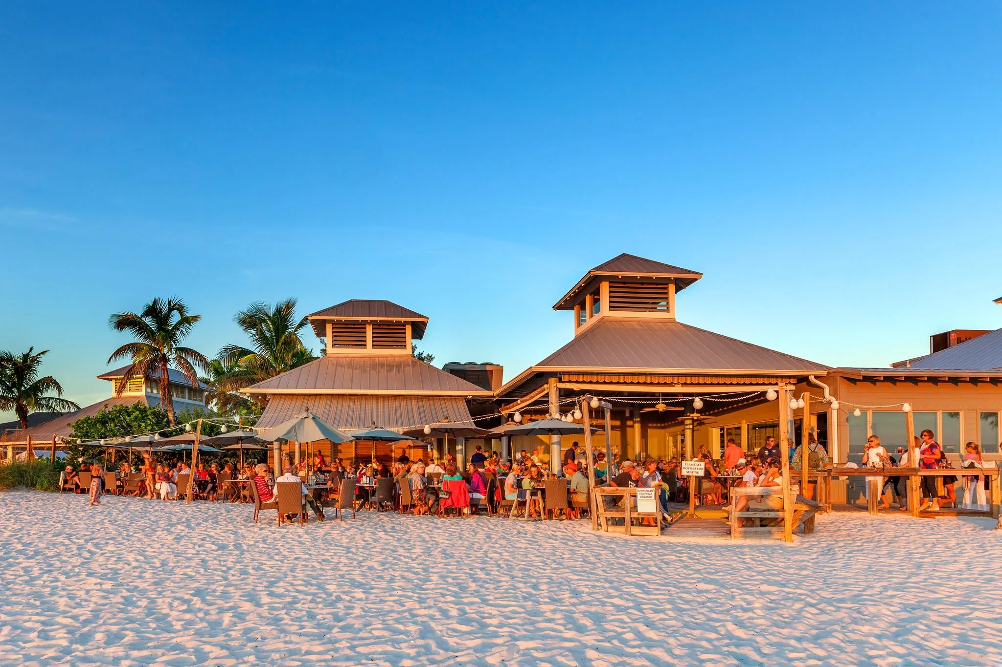 Sarasota Motor Club - Premium Car Service Sarasota - Beachside restaurant with many people dining under umbrellas and string lights, surrounded by palm trees at sunset.