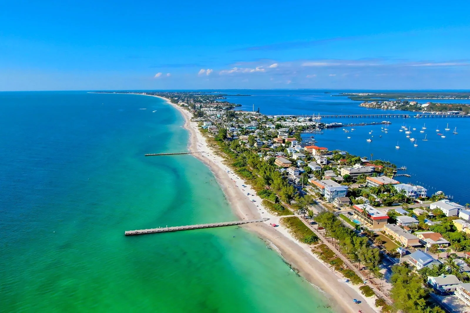 Sarasota Motor Club - Premium Car Service Sarasota - Aerial view of a coastal town with a sandy beach and turquoise ocean on the left, and a waterfront with houses, boats, and a marina on the right under a blue sky.