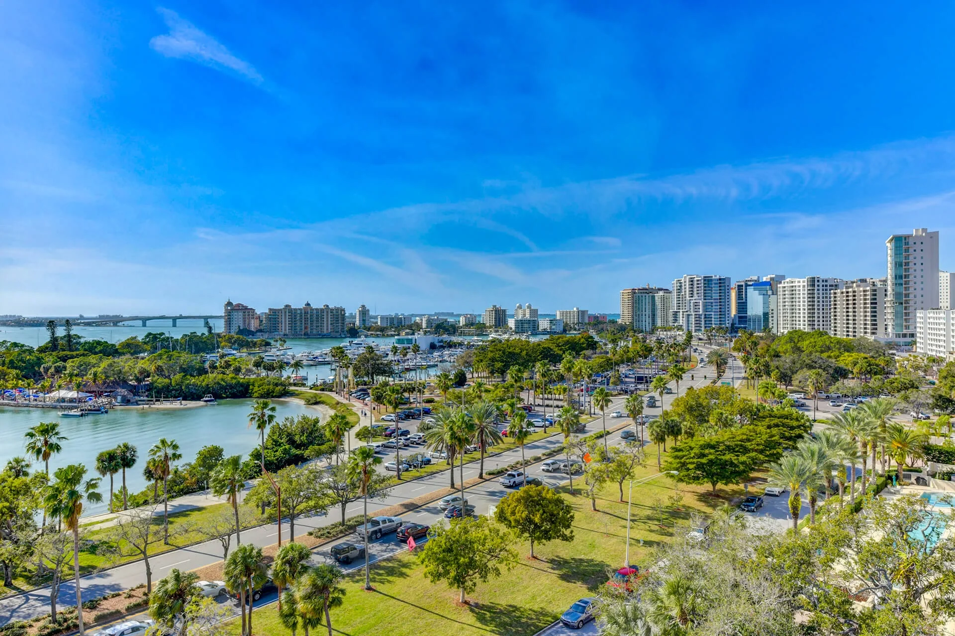 Sarasota Motor Club - Premium Car Service Sarasota - A panoramic view of a city skyline with tall modern buildings, a waterfront with boats, and a park with palm trees and parking lots under a clear blue sky.