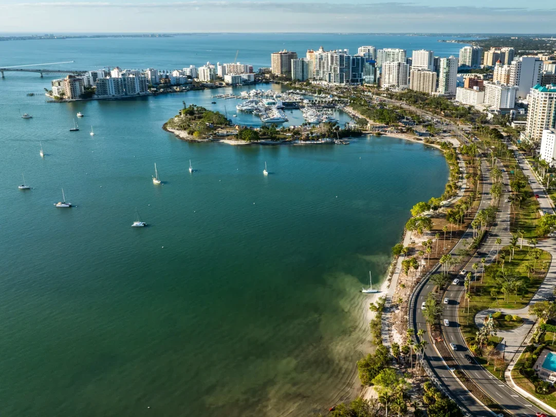Sarasota Motor Club - Exotic Car Consignment Sarasota - Aerial view of a city shoreline with high-rise buildings, marina with boats, and a curved road lined with palm trees along the water.
