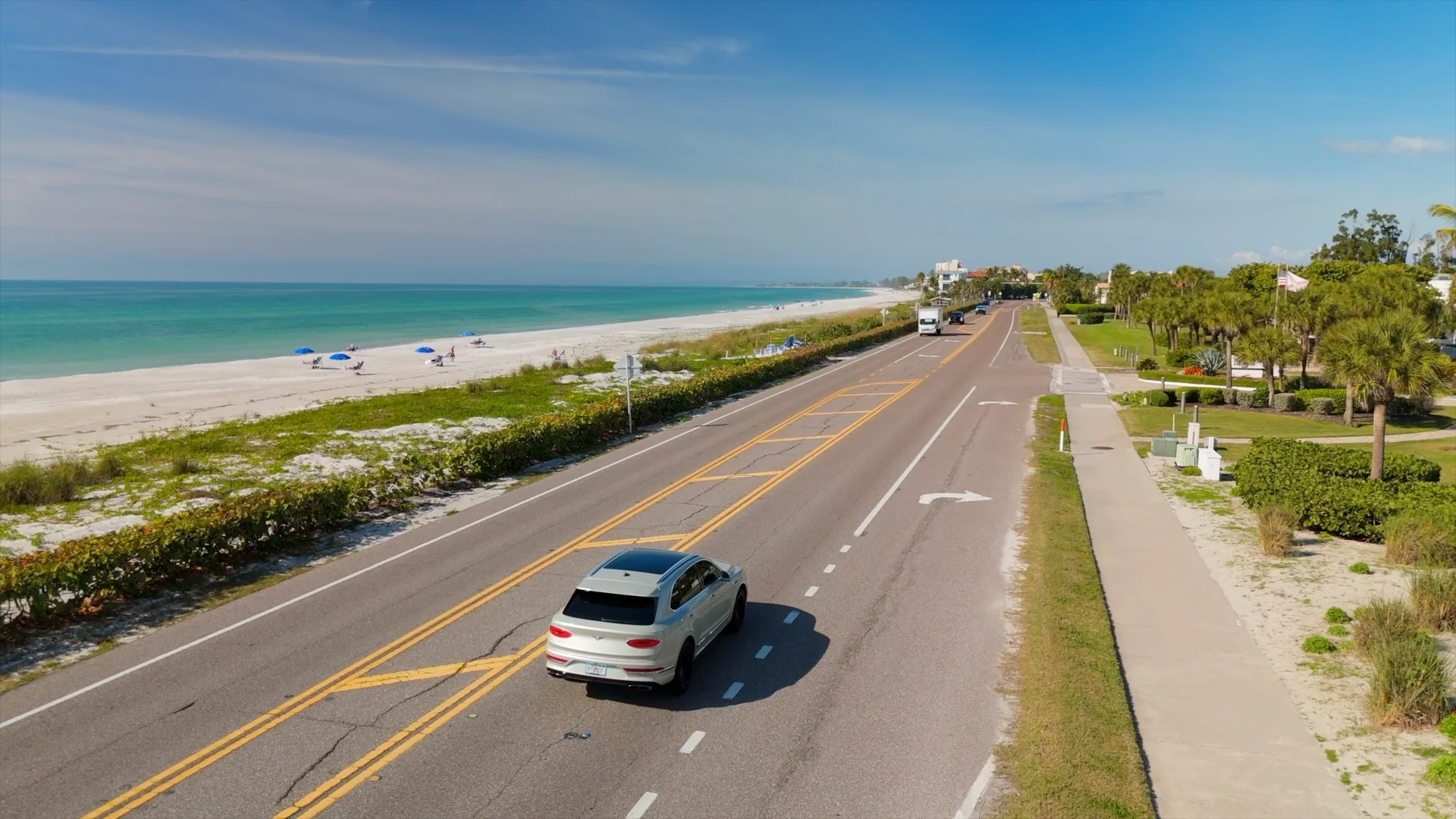 Sarasota Motor Club -  luxury car rental sarasota - A scenic beachside road with a silver Bentley driving along, lined with lush palm trees and greenery on the right, and a sandy beach with blue umbrellas and people sunbathing on the left.