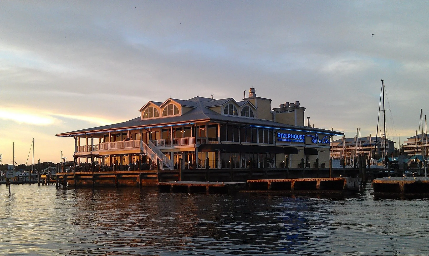 Sarasota Motor Club - Exotic Car Rental Sarasota Florida - Waterfront restaurant with a large balcony, several windows, and a neon sign that says "RIVERHOUSE" in a marina with sailboats, under a cloudy sky at sunset.