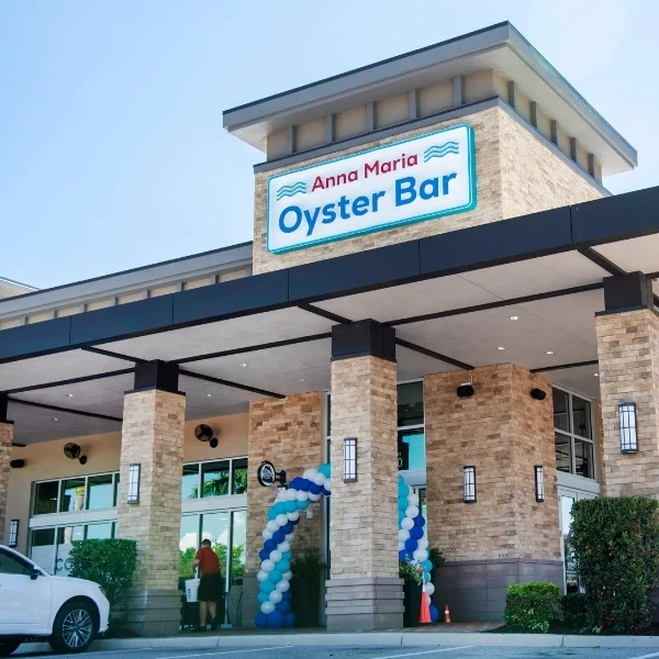 Sarasota Motor Club - Exotic Car Rental Sarasota Florida - Exterior view of Anna Maria Oyster Bar with a sign above the entrance, decorated with blue, white, and light blue balloons, and a person walking in front of the building.