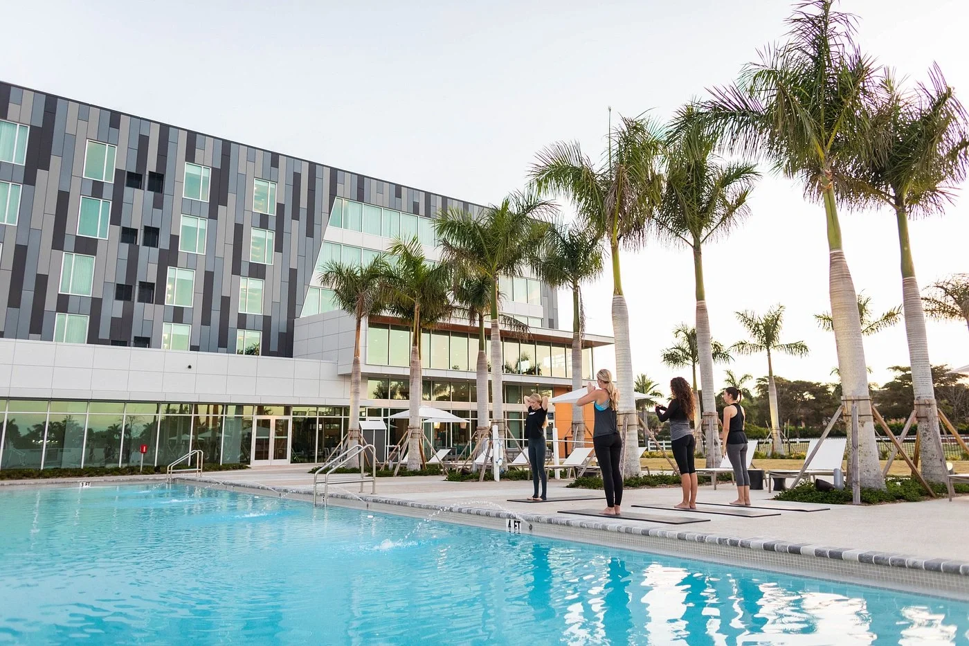 Sarasota Motor Club - Exotic Car Rental Sarasota Florida - Four women doing yoga by a swimming pool at a modern hotel with tall palm trees.