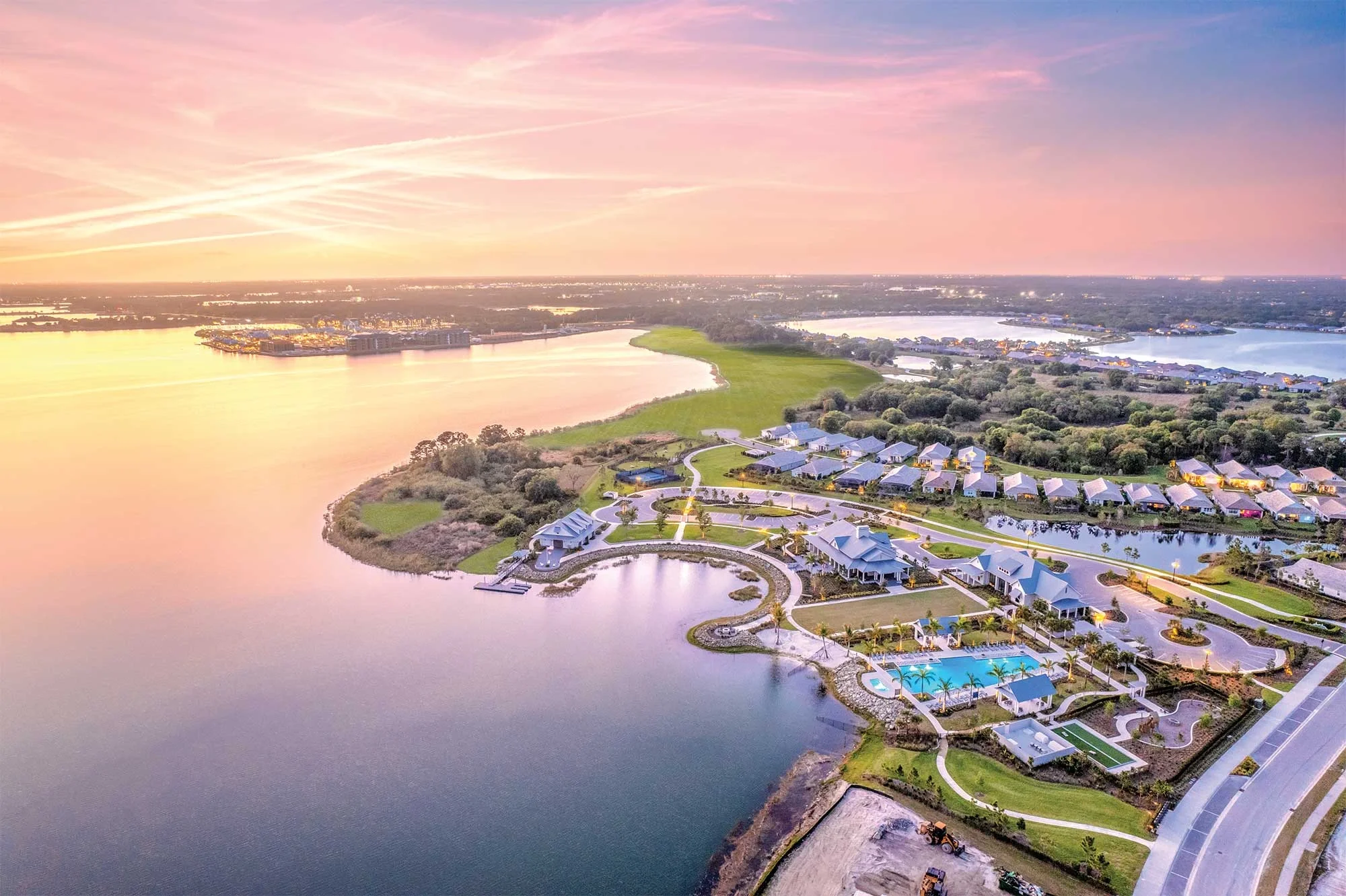 Sarasota Motor Club - Exotic Car Consignment Sarasota - Aerial view of a lakeside residential community at sunset with houses, a swimming pool, green lawns, and winding roads.