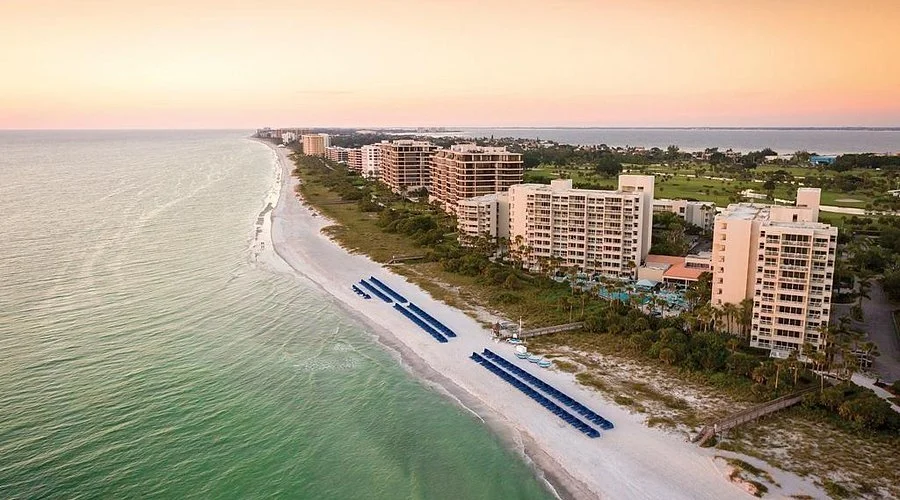 Aerial view of beachfront resort with multiple hotels and condominium buildings along a sandy coast at sunset, with palm trees and green vegetation near the shore.