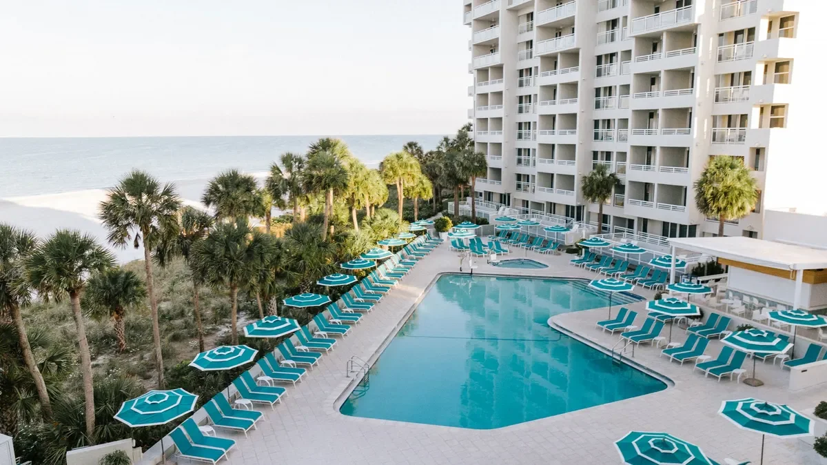 Swimming pool area with blue and white striped umbrellas and lounge chairs, multiple stories hotel building, and palm trees, near the beach and ocean.