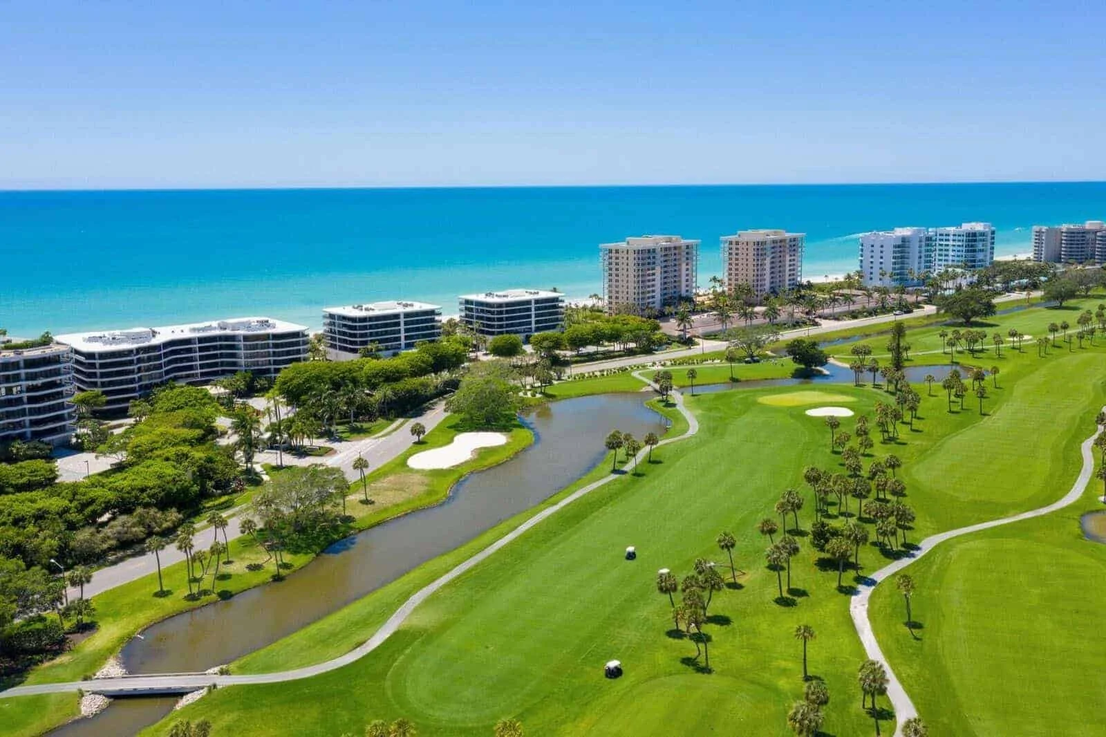 Aerial view of a coastal golf course with lush green fairways, sand bunkers, and small ponds, adjacent to high-rise beachfront buildings and the ocean in the background.