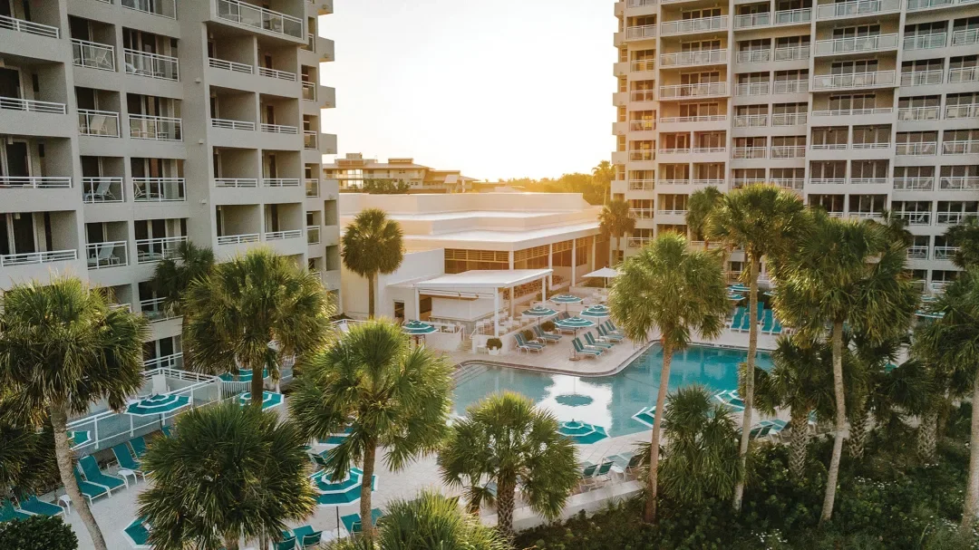 View of a swimming pool surrounded by palm trees, lounge chairs, and tall apartment buildings at a resort or hotel during sunset.