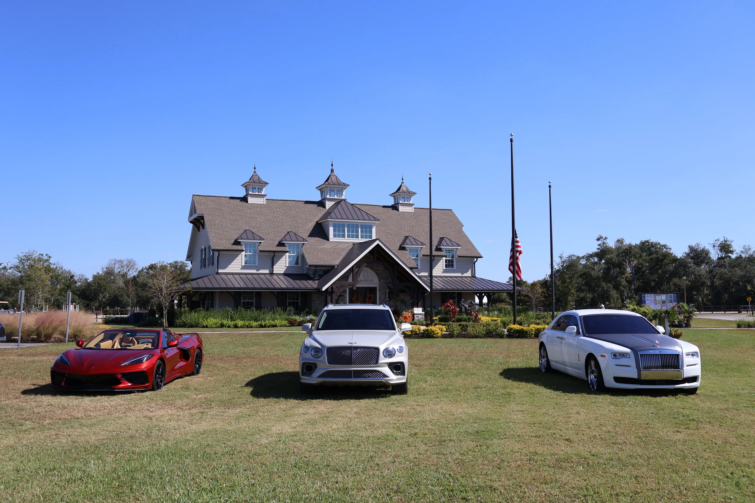 Sarasota Motor Club - sports car rental sarasota fl - A large, elegant house with a landscaped front yard, three luxury cars parked on the grass in front of the house, American flags, and a bright blue sky.