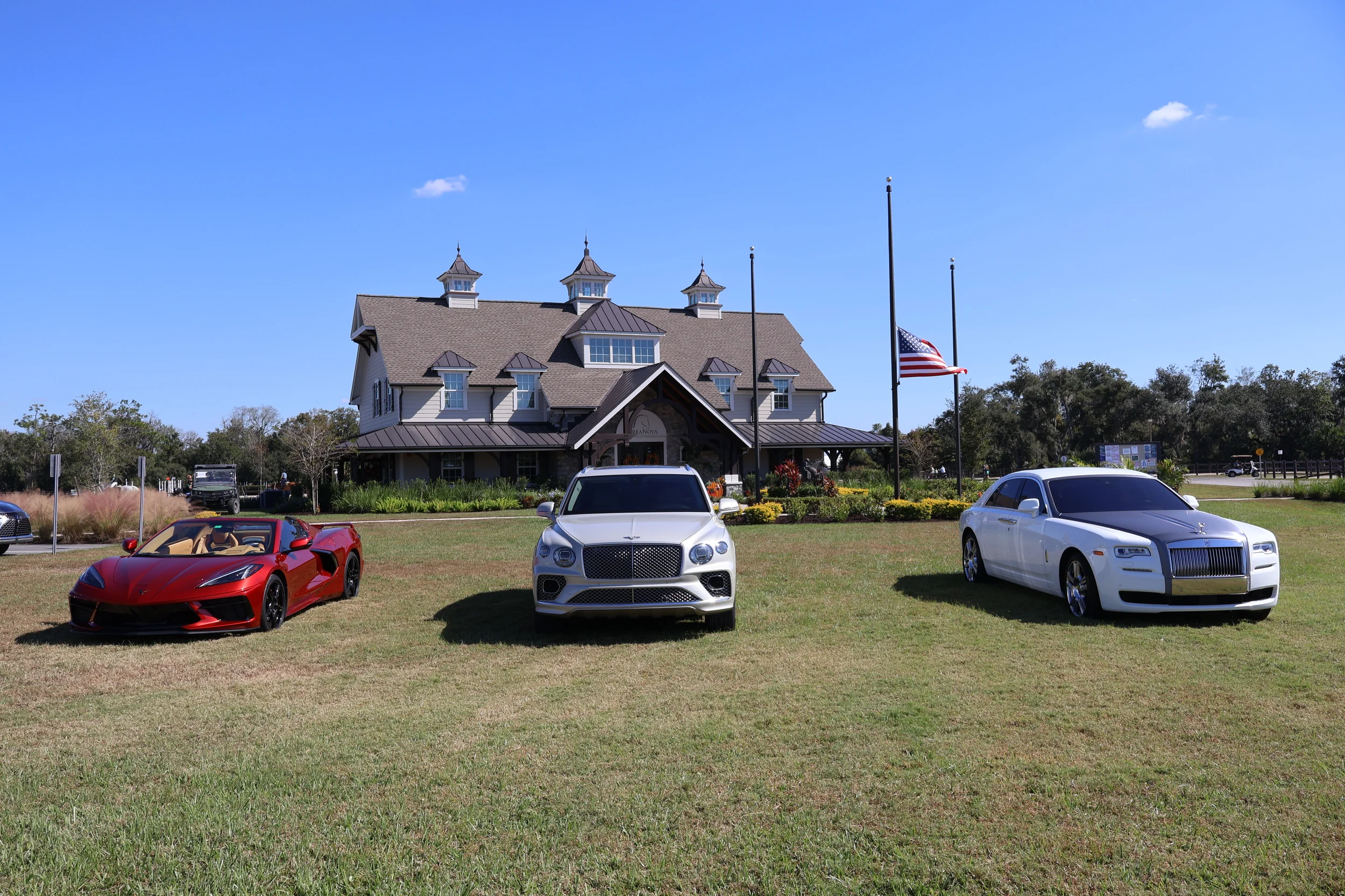 Sarasota Motor Club - sports car rental sarasota fl - Three luxury cars parked on grass in front of a large, house-like building with American flags, a landscaped garden, and trees, under a blue sky.