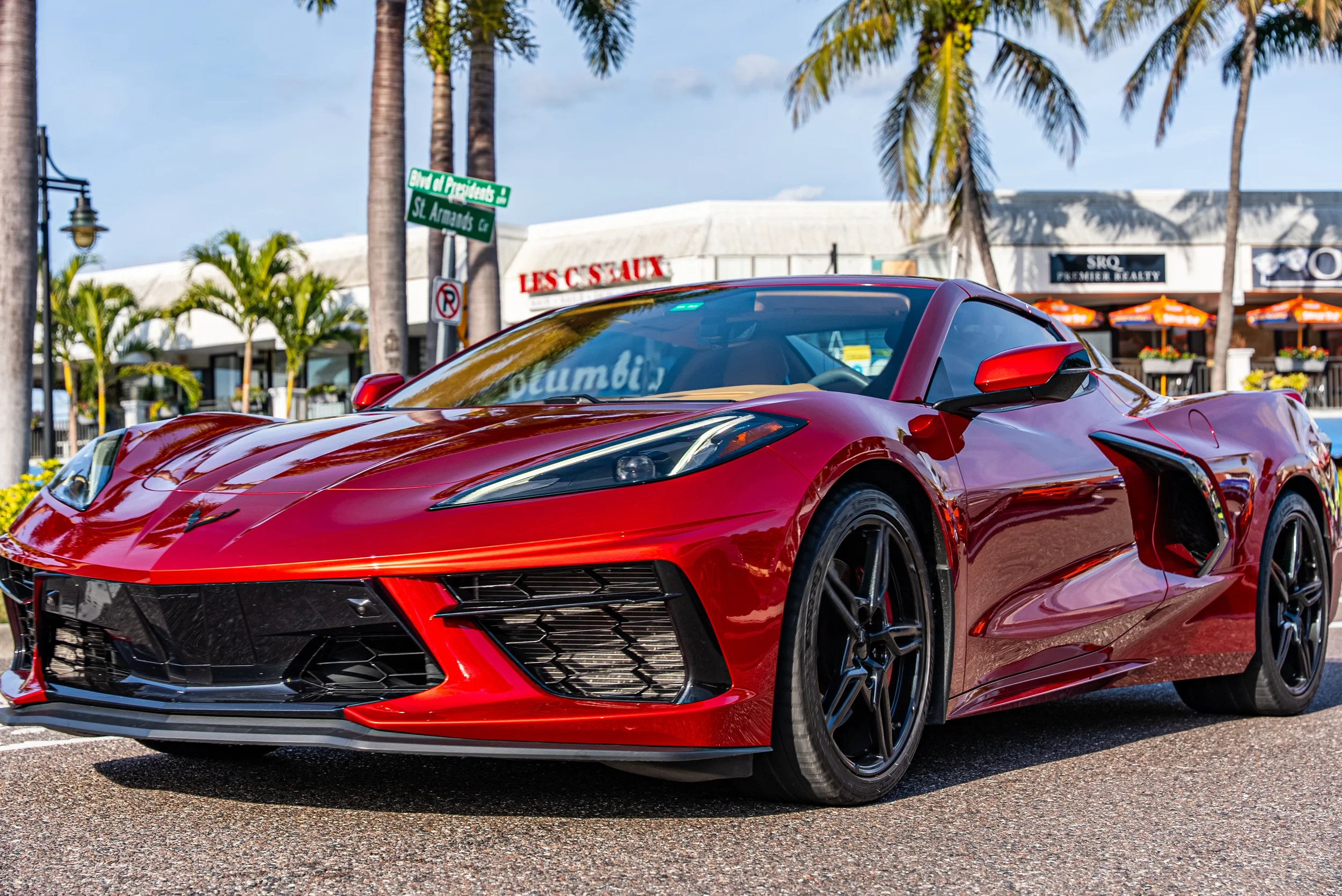 Sarasota Motor Club - Corvette rental Longboat Key - A red sports car parked on a street with palm trees and shopping stores in the background.